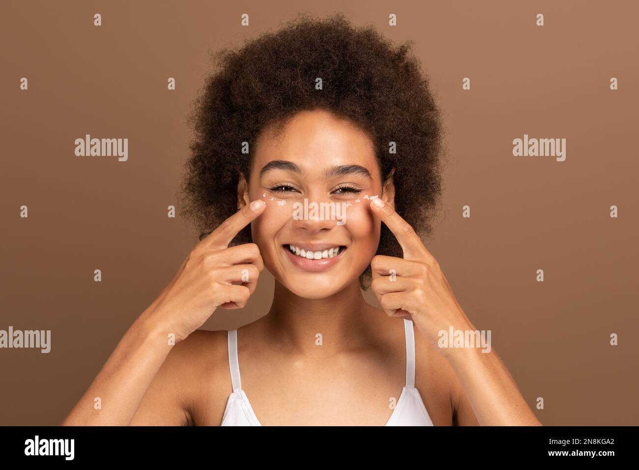 Gaie jeune afro-américaine curly dame avec la peau parfaite en blanc dessus appliquer la crème sur le visage sous les yeux Banque D'Images