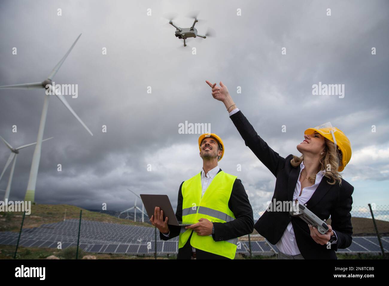 Deux ingénieurs au travail font voler un drone et utilisent l'ordinateur portable dans une ferme solaire avec des éoliennes Banque D'Images