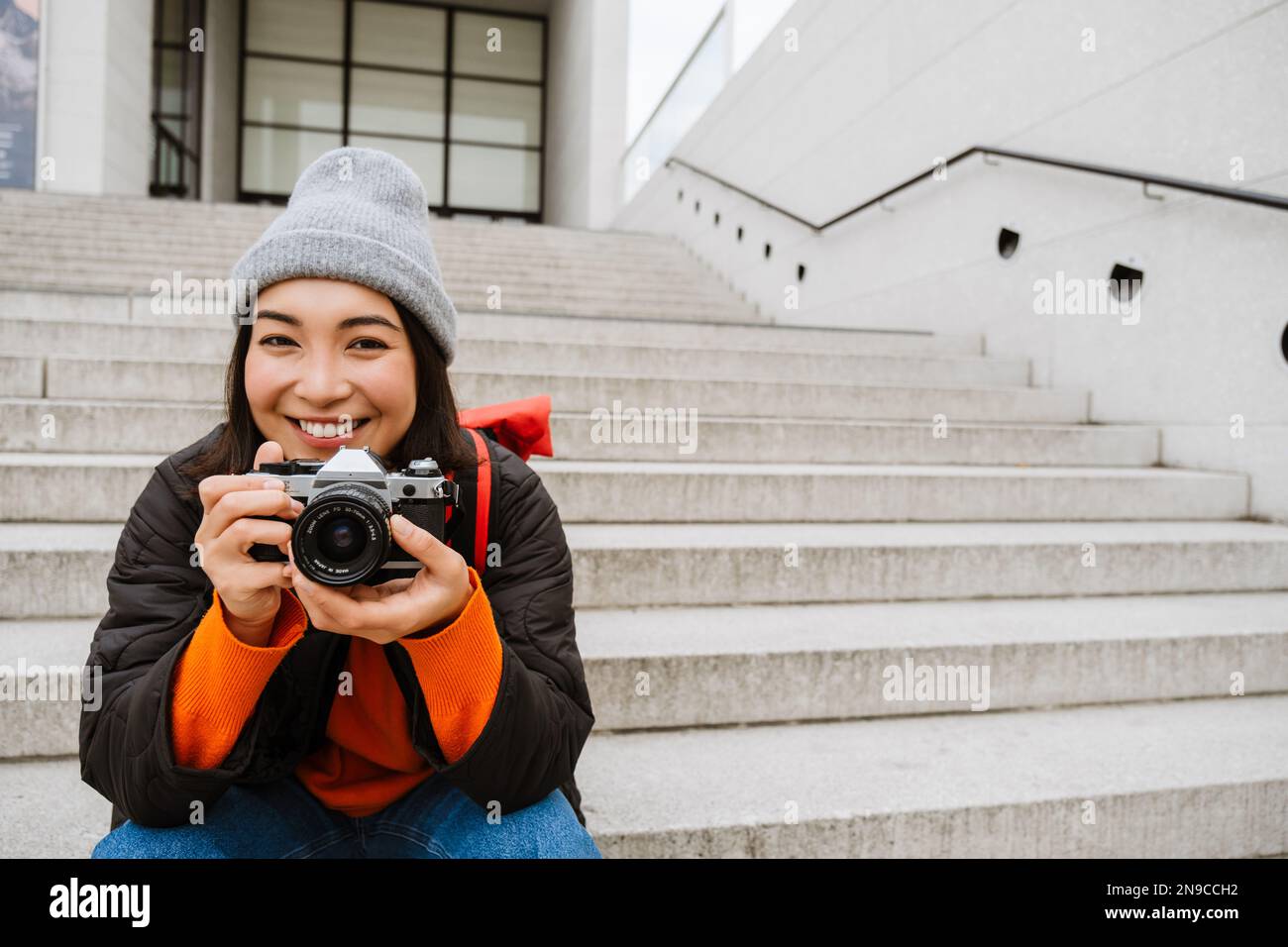 Belle jeune femme asiatique souriante dans des vêtements chauds prendre des photos avec un appareil photo d'époque tout en étant assise sur les escaliers dans la rue de la ville Banque D'Images