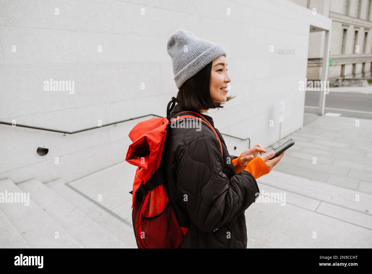 Belle jeune femme asiatique souriante avec sac à dos en utilisant un smartphone tout en se tenant sur les escaliers dans la rue de la ville Banque D'Images
