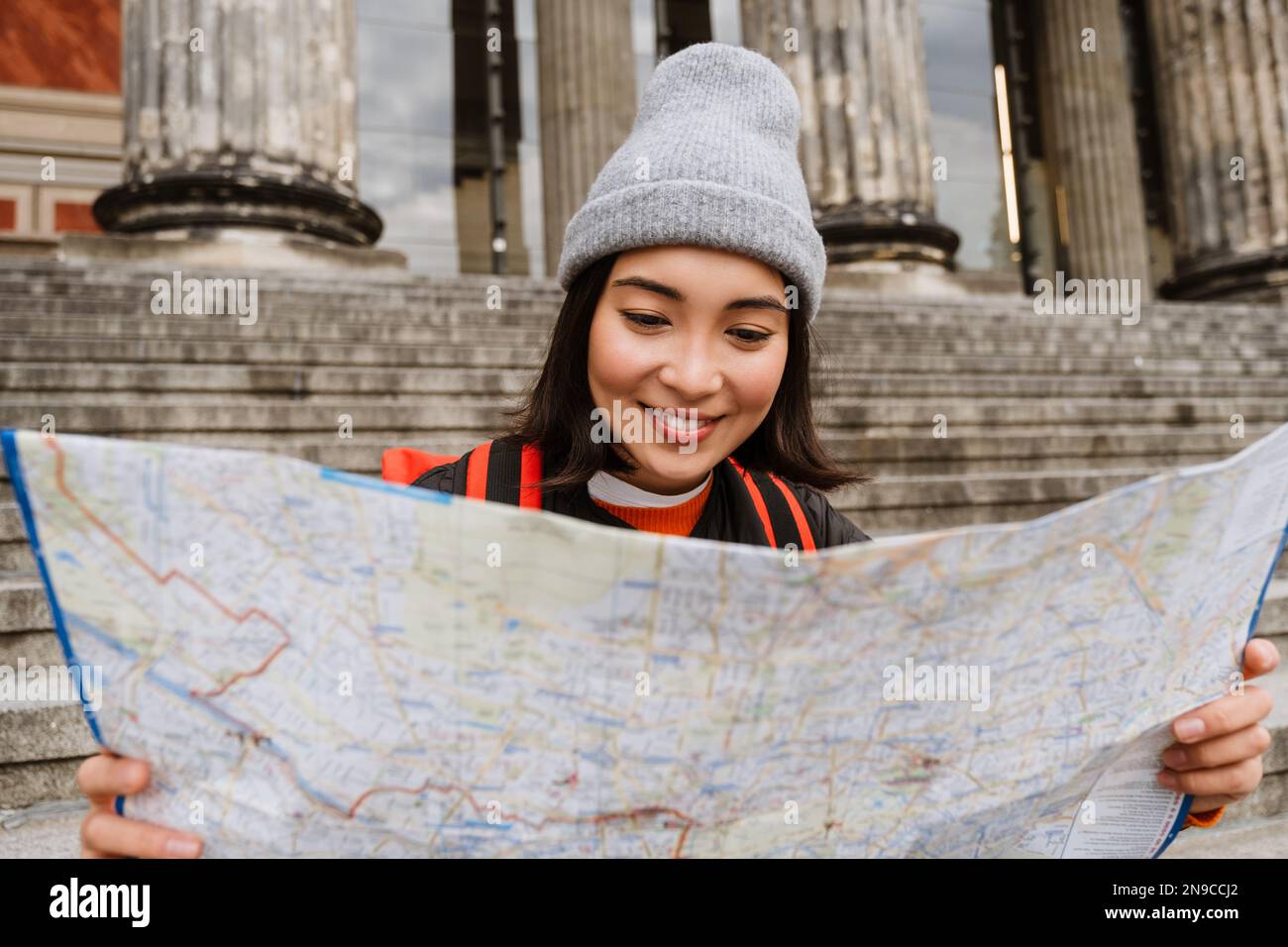 Jeune femme asiatique joyeuse touriste avec sac à dos examinant la carte papier tout en étant assis sur les escaliers dans la vieille ville rue Banque D'Images