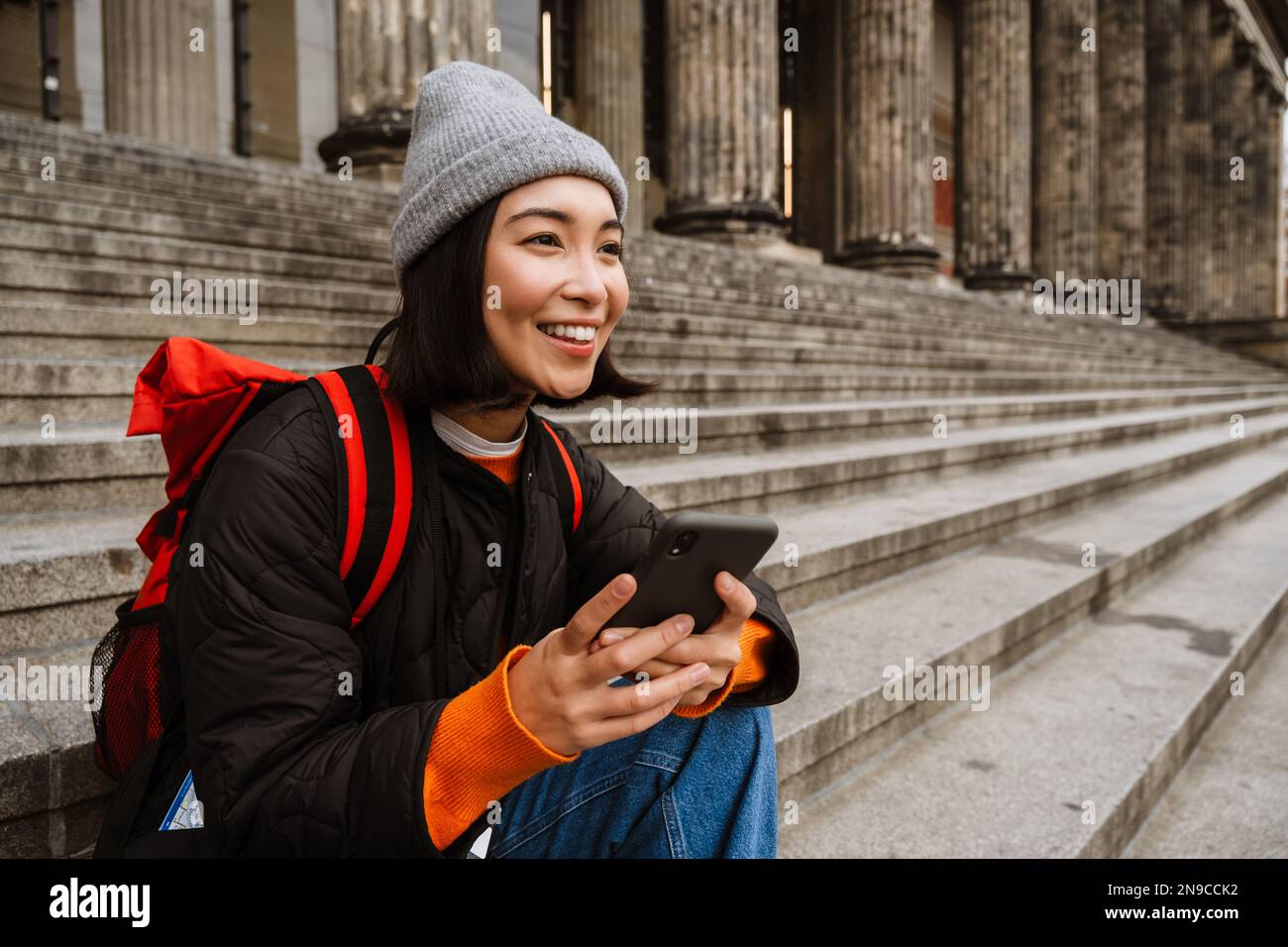 Belle jeune femme asiatique souriante avec sac à dos en utilisant un smartphone tout en étant assis sur les escaliers dans la vieille ville rue Banque D'Images
