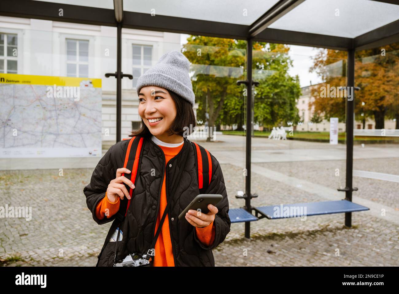 Jeune femme asiatique gaie portant des vêtements chauds à l'aide d'un smartphone et regardant de côté en se tenant à la gare routière Banque D'Images