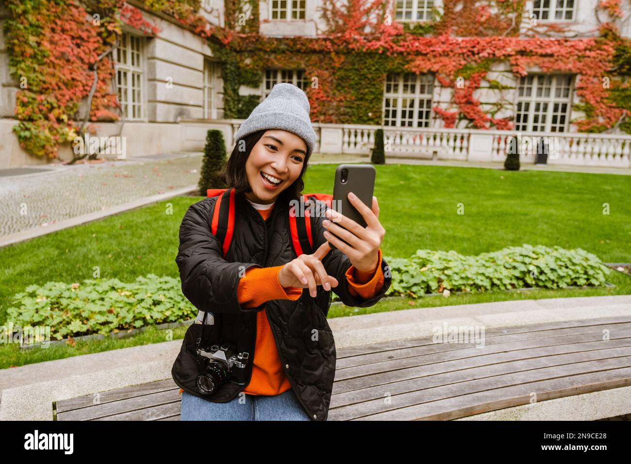 Belle jeune femme asiatique souriante vêtue de vêtements chauds emportant un selfie sur un téléphone portable tout en étant assise sur un banc dans la vieille ville Banque D'Images