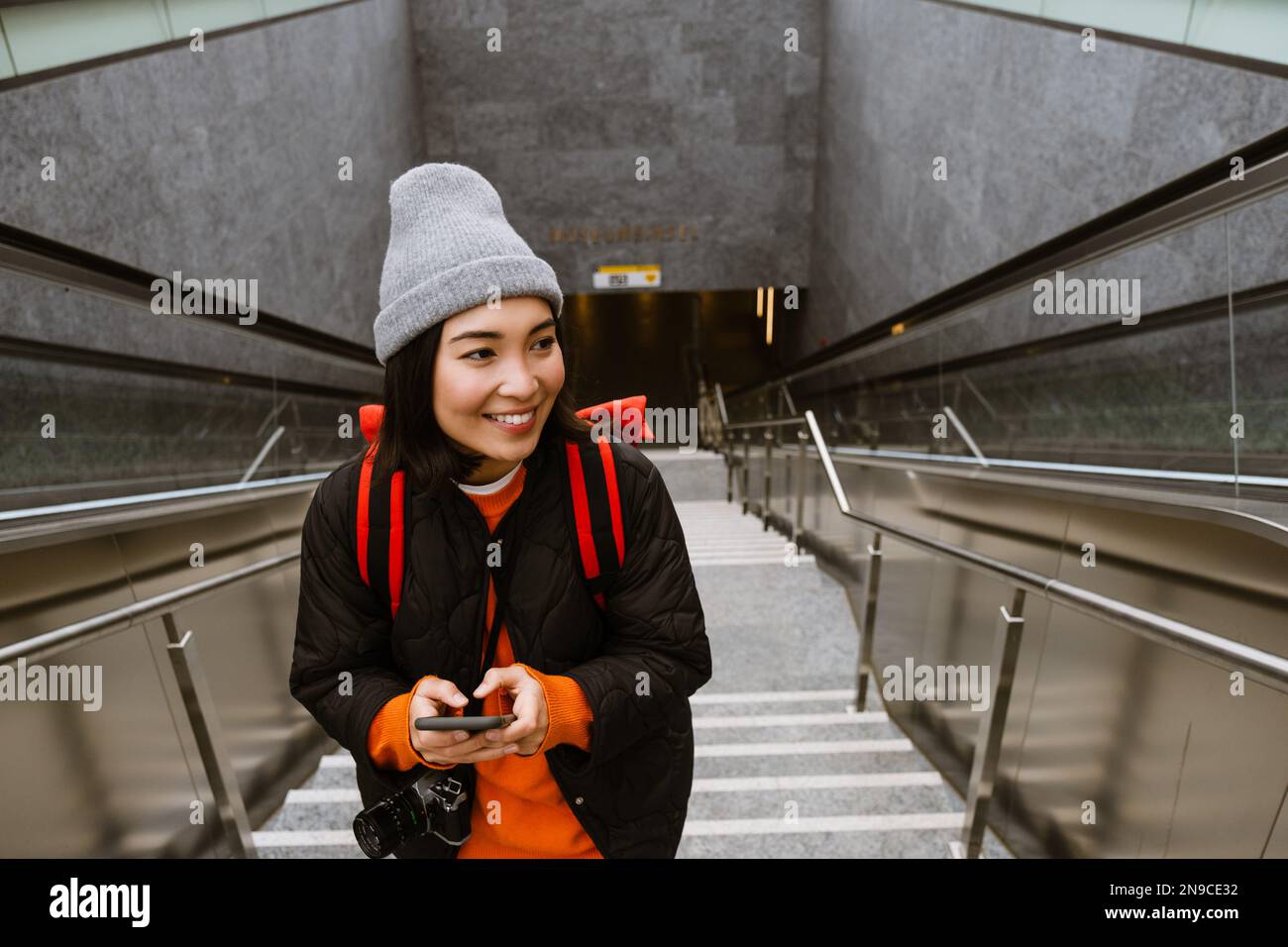Belle jeune femme asiatique souriante dans des vêtements chauds en utilisant un smartphone tout en montant les escaliers à l'extérieur Banque D'Images