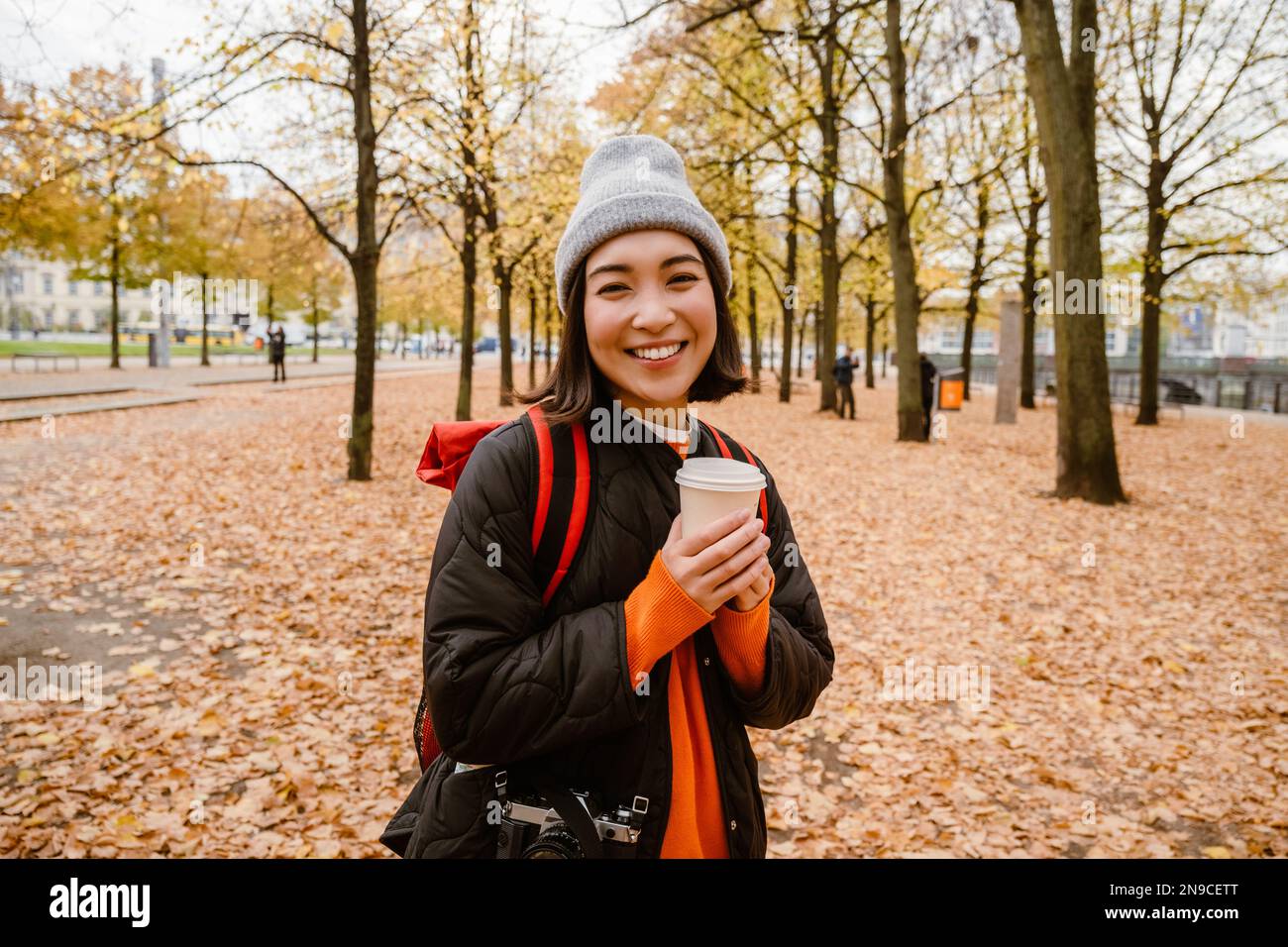 Belle jeune femme asiatique souriante portant des vêtements chauds buvant un café à emporter pendant la promenade dans le parc d'automne Banque D'Images