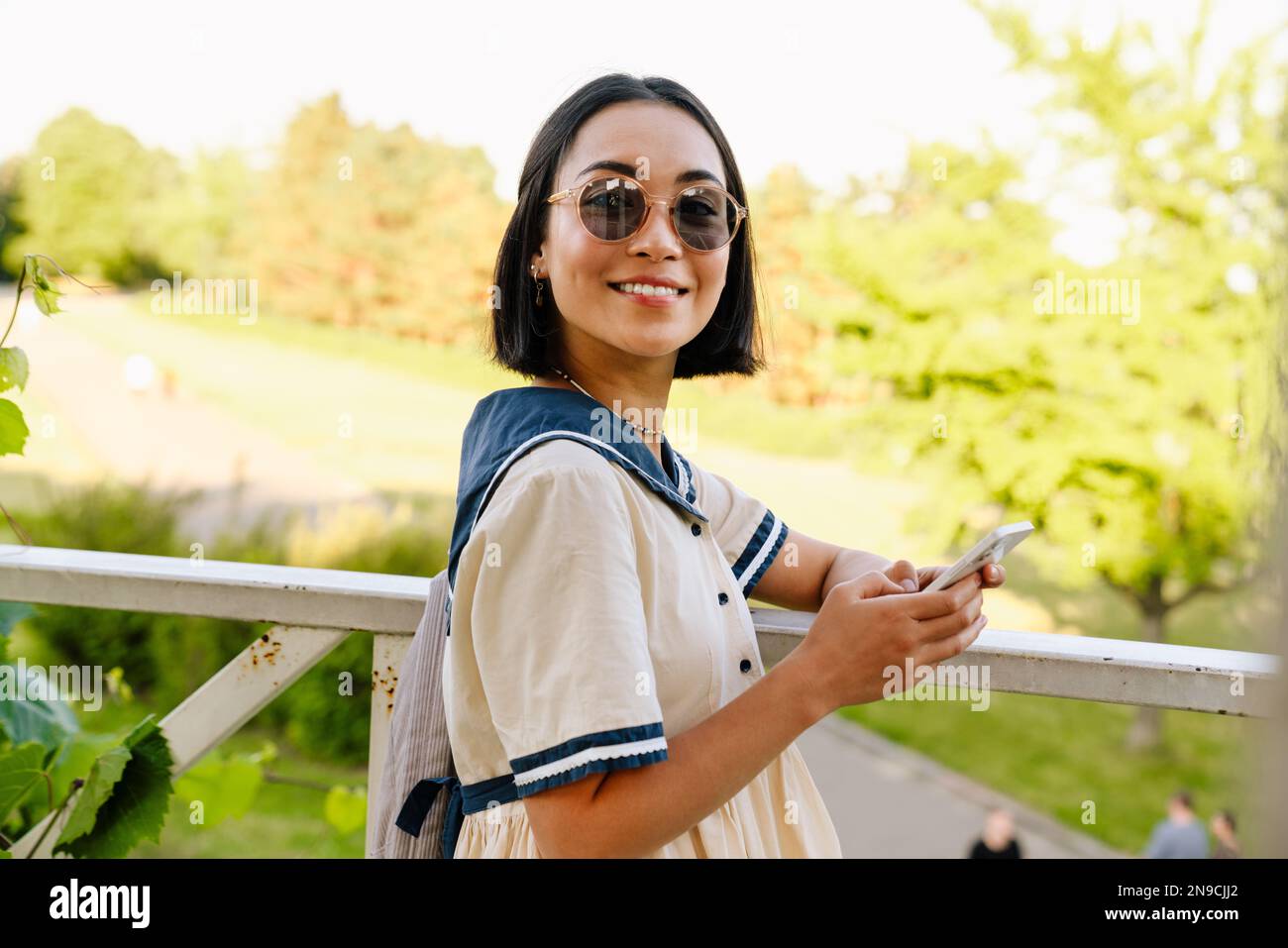 Jeune belle fille asiatique en lunettes de soleil souriant et en utilisant  le téléphone mobile tout en se tenant dans le parc vert Photo Stock - Alamy