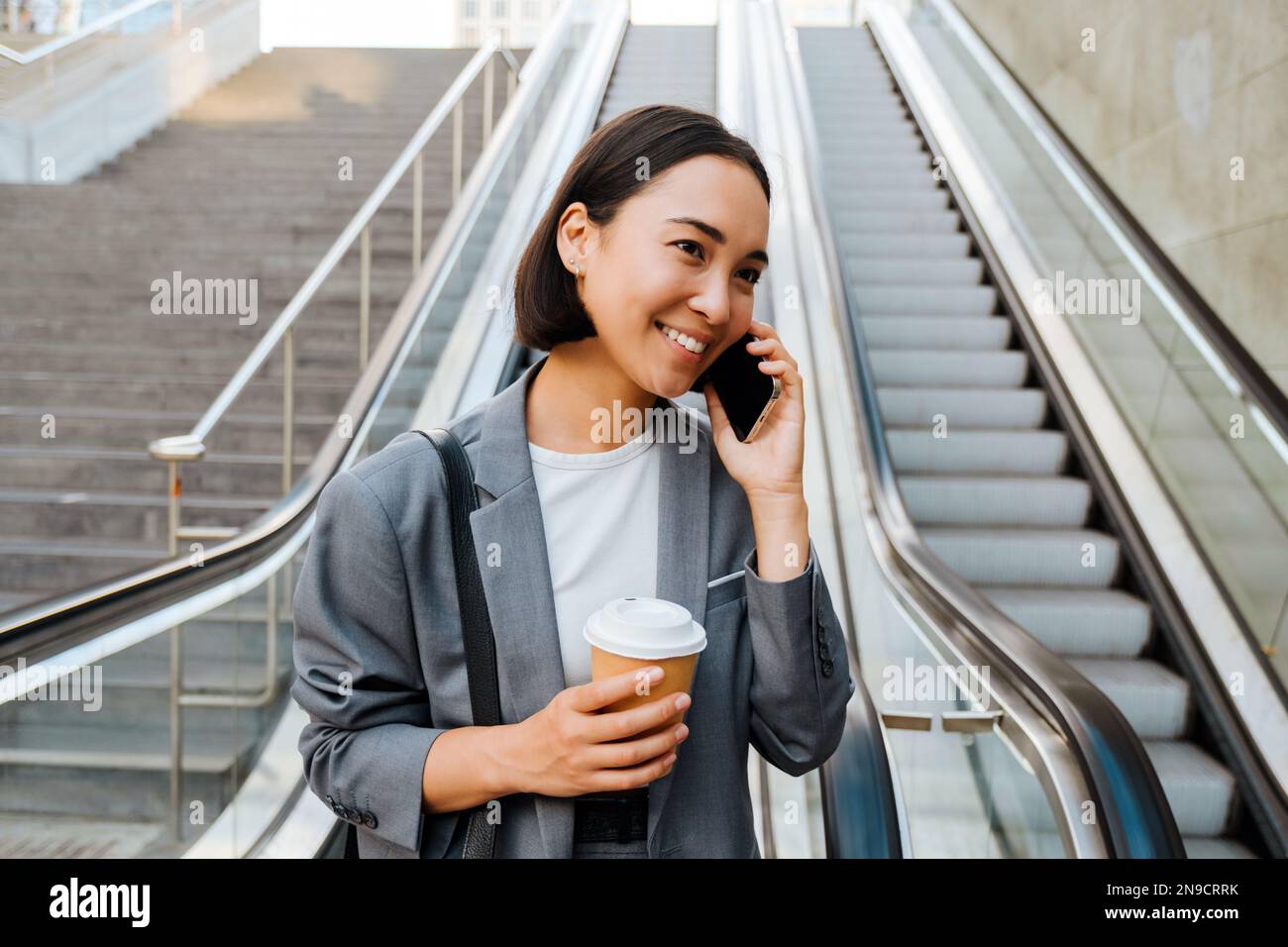 Jeune femme asiatique parlant au téléphone portable et tenant le café tout en descendant l'escalier roulant à l'extérieur Banque D'Images