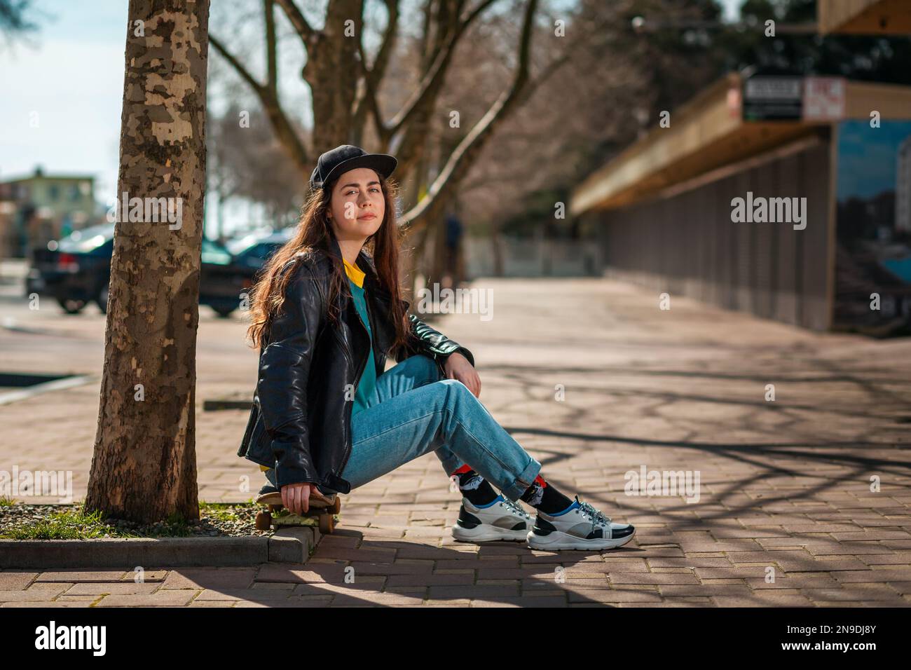 Portrait d'une belle fille de l'adolescence d'hipster dans une veste en  cuir assis sur un skateboard. Vue latérale. Il y a une allée avec des  arbres à l'arrière.concept de s Photo Stock -