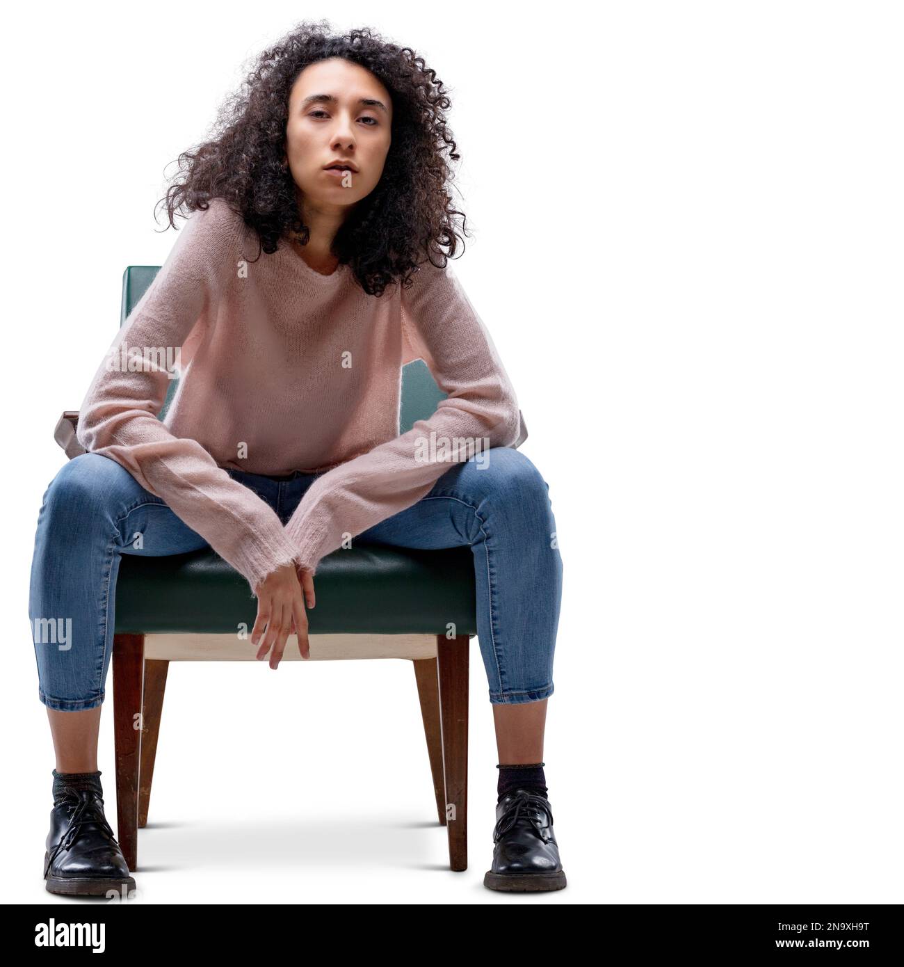 Jeune femme aux cheveux mauris assise sur une chaise avec des jambes écartées dans une position de tomboy et une expression de confiance et de défi sérieux. Isolé sur Banque D'Images
