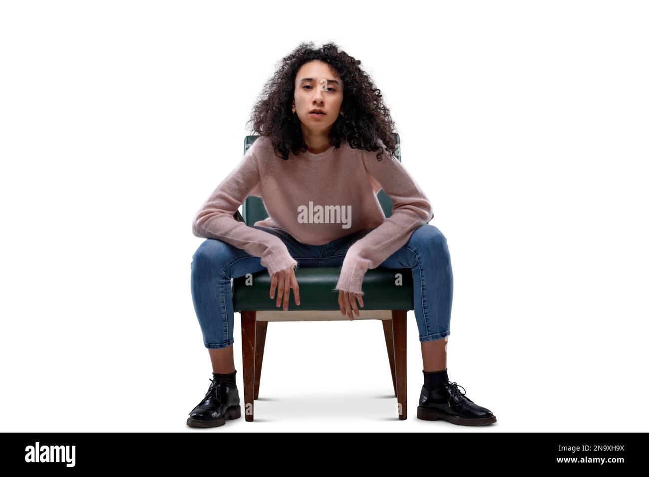 Jeune femme aux cheveux bouclés assise sur une chaise dans une position gaieuse et provocante, coudes reposant sur les cuisses. Elle vous défie. Isolé sur transparent ou wh Banque D'Images