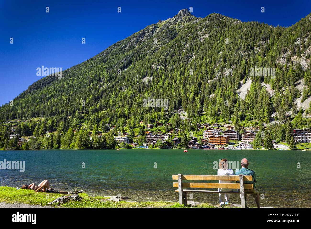 Un couple profite de la vue sur un banc au bord du lac de Champex sous ciel bleu, montagne et stations sont en arrière-plan ; Champex, Valais, Suisse Banque D'Images