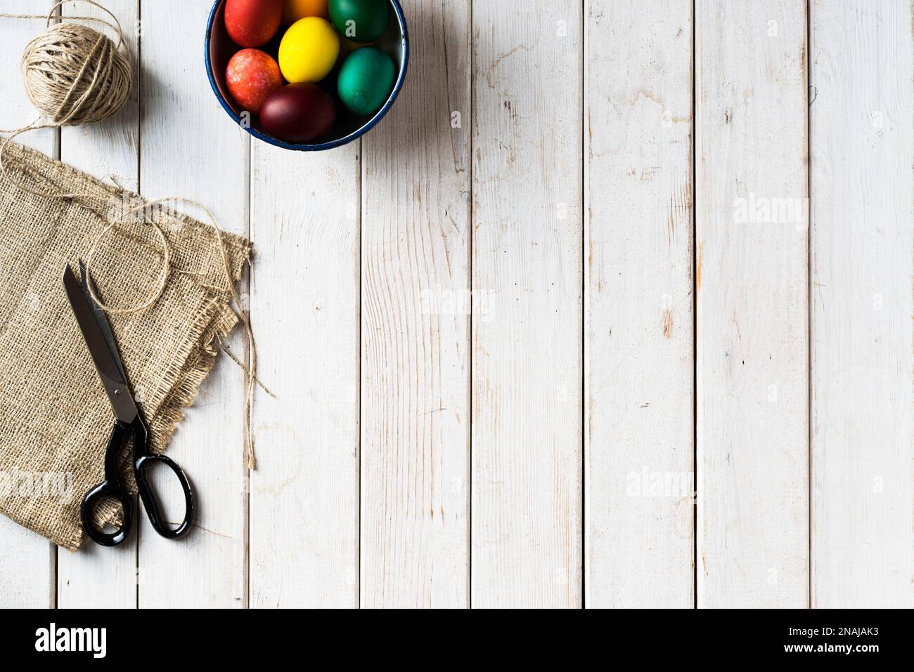 Arc-en-ciel couleur oeufs de pâques dans un bol. Ciseaux, toile de jute, corde sur fond en bois. Directement au-dessus du plan d'examen. Banque D'Images