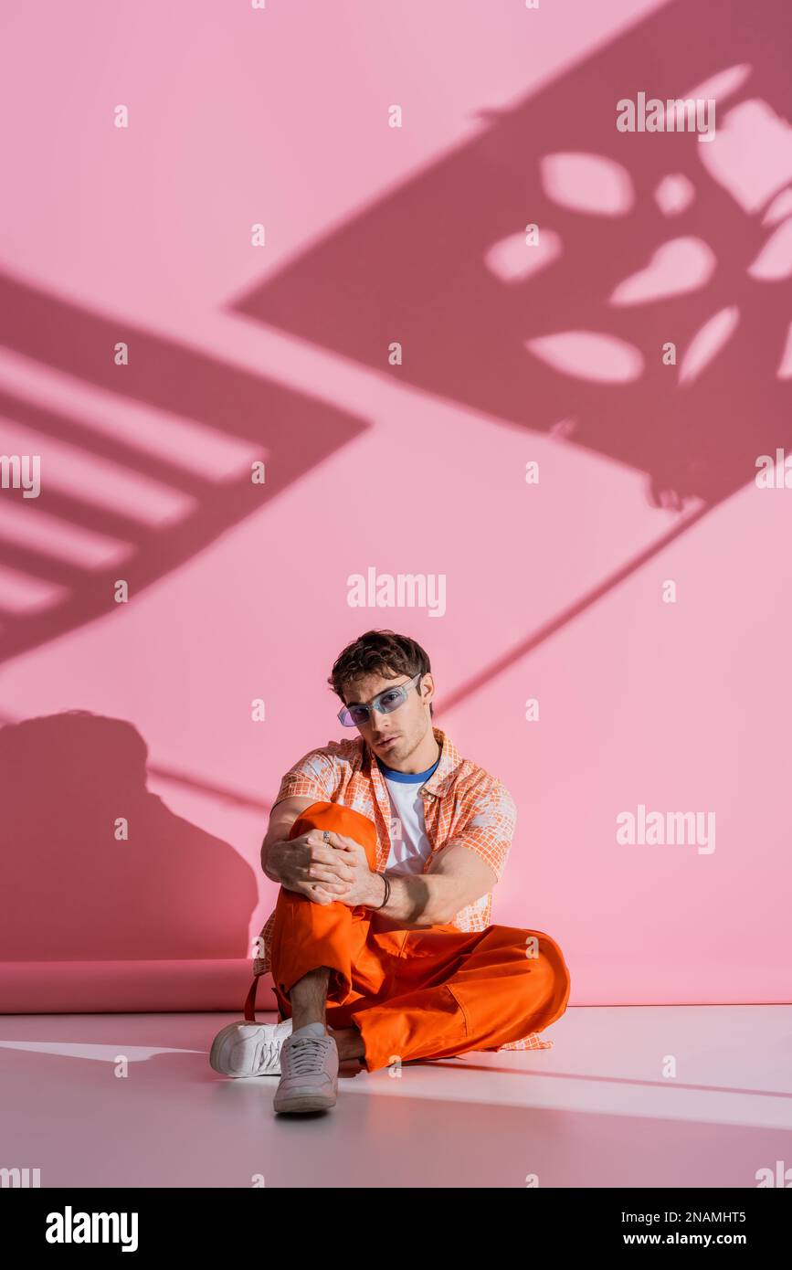 Jeune homme tendance en lunettes de soleil assis sur fond rose avec ombre, image de stock Banque D'Images