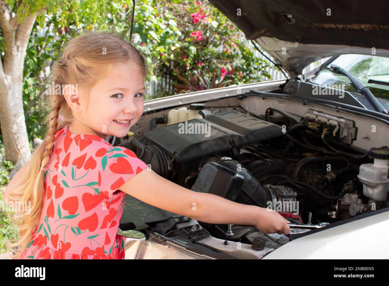 Petite fille soupirante caucasienne fixant le moteur d'une voiture avec un capot de voiture ouvert Banque D'Images