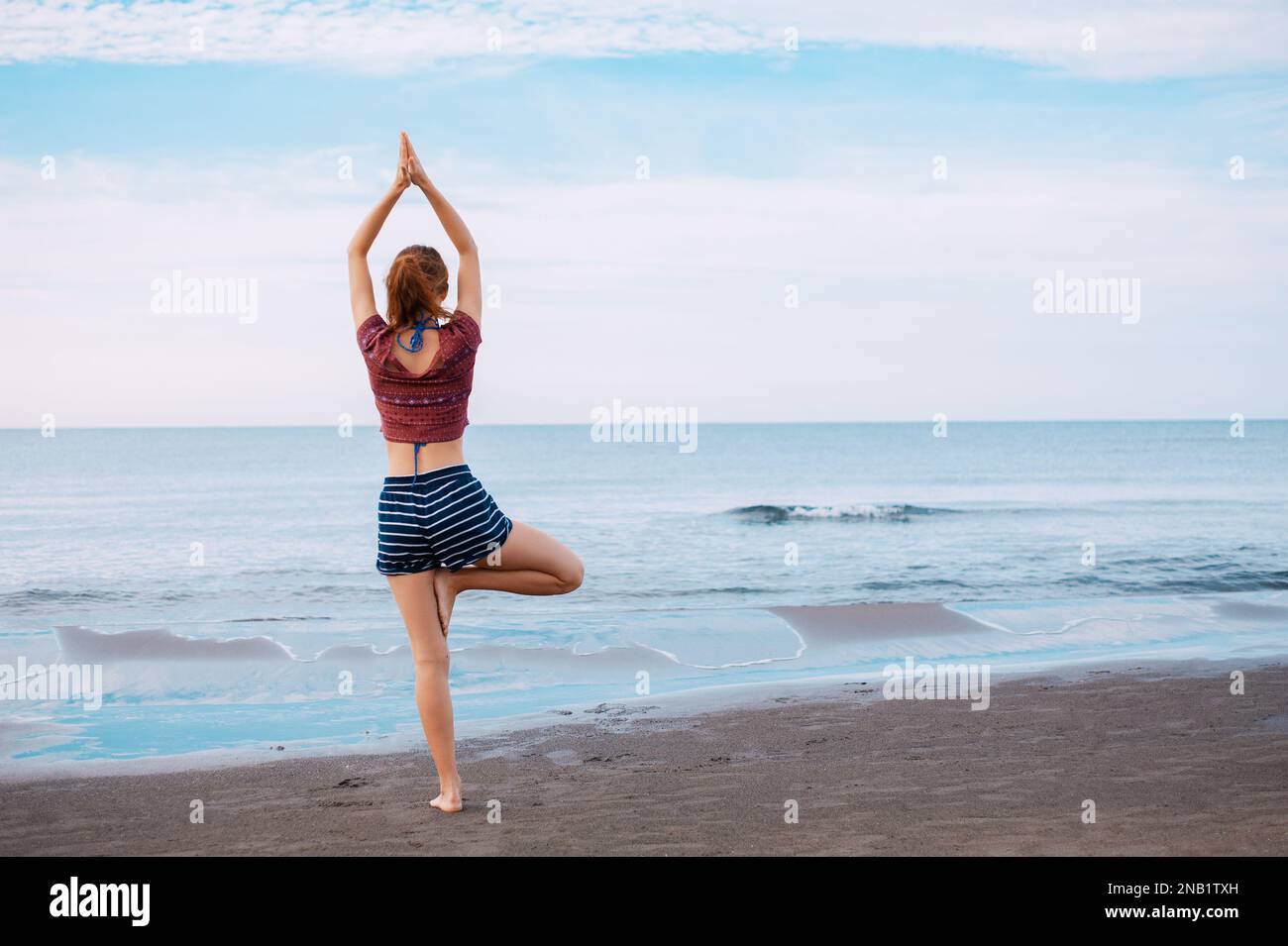 Vue arrière de la jeune femme à tête rouge faisant de l'exercice de yoga à vrikssasana pose sur la rive de la plage au Monténégro Ulcinj, paisible scène mer Adriatique Banque D'Images