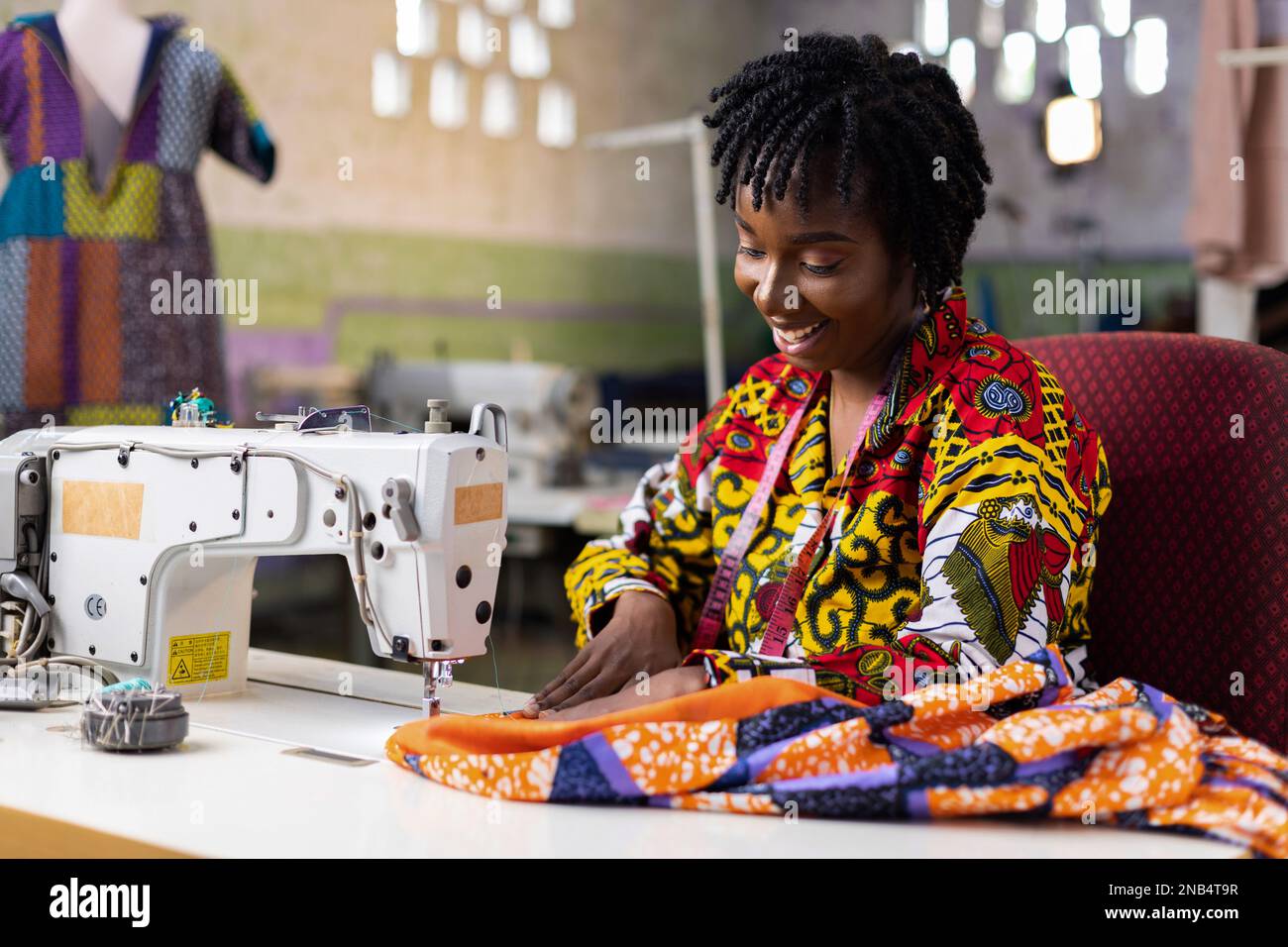 Femme africaine de mode tailleur portant une robe ankara avec des cheveux locs coudre une tenue dans son studio de couture avec une machine électrique. Banque D'Images