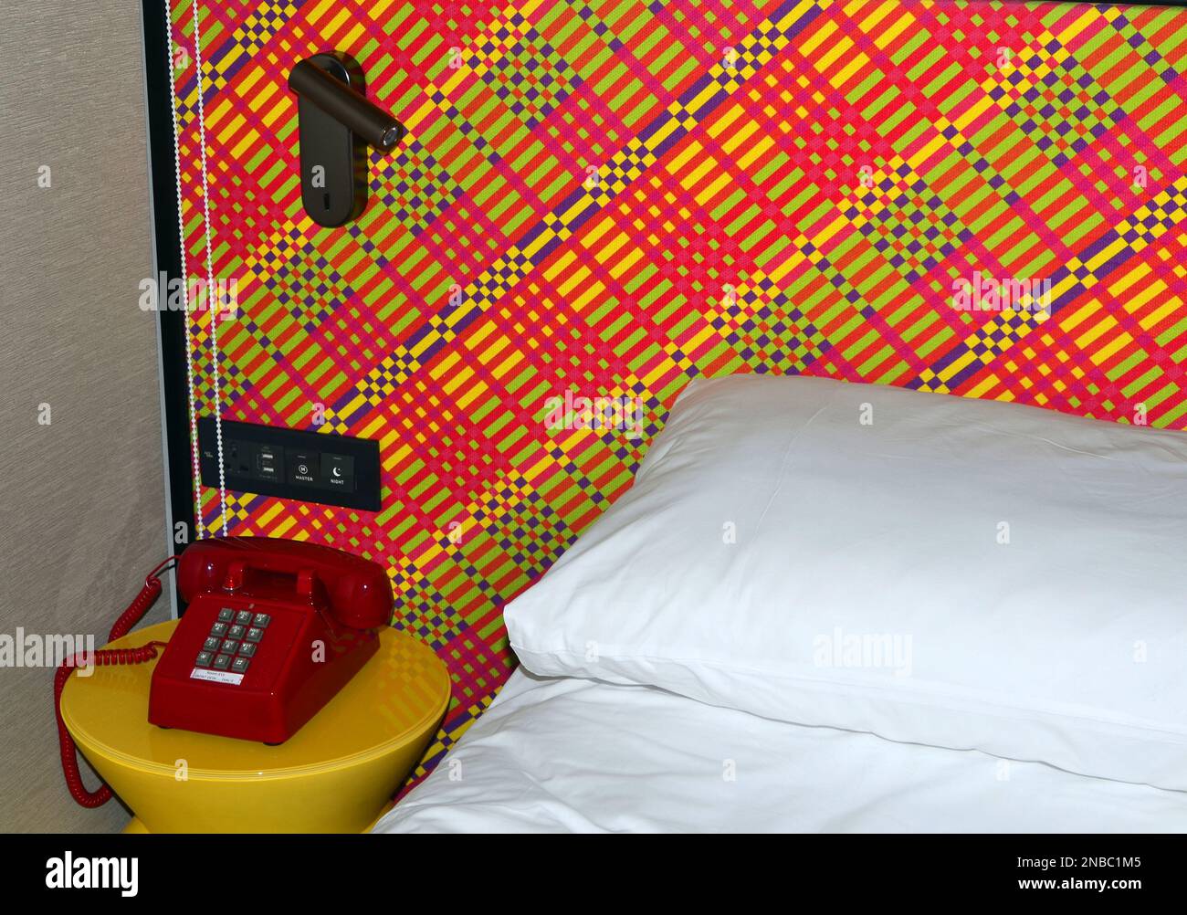Un téléphone ou un téléphone rouge vif sur une table jaune vif à côté du coin d'un lit avec une feuille blanche et taie d'oreiller; mur a un design multicolore. Banque D'Images