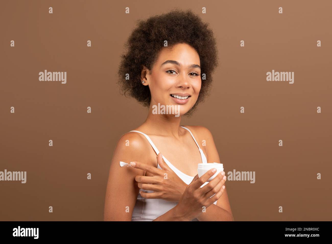 Gaie jeune afro-américaine cally dame avec la peau parfaite dans blanc top tenir le pot, appliquant de la crème sur l'épaule Banque D'Images