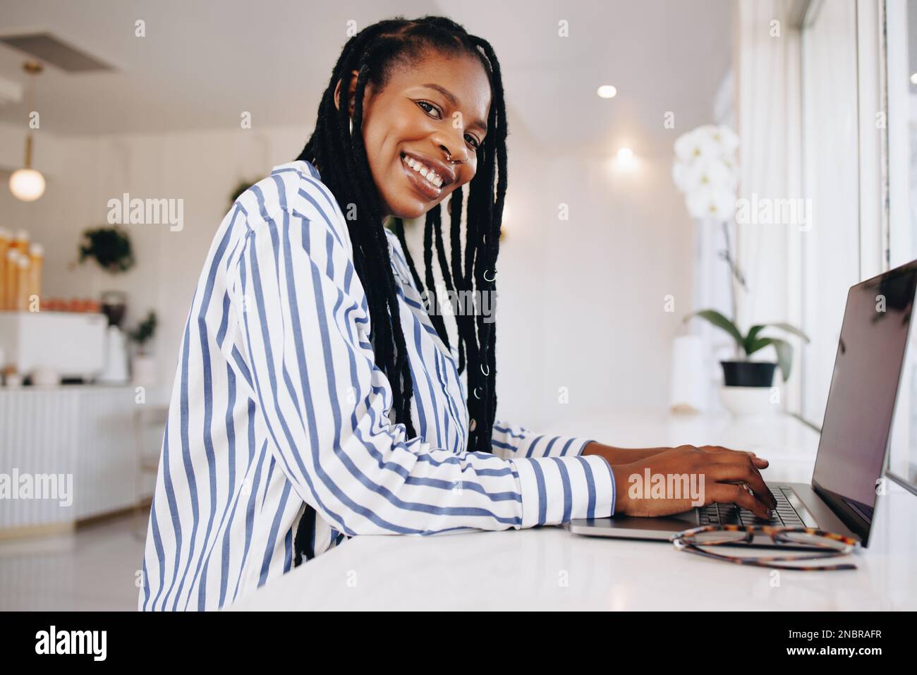 Une femme joyeuse, pigiste en souriant à la caméra tout en tapant sur un ordinateur portable dans un café. Bonne jeune femme d'affaires noire travaillant en ligne dans un café. Banque D'Images