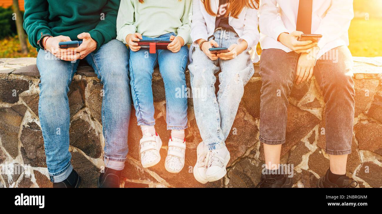 Quatre enfants qui naviguent sur leur smartphone tout en étant assis sur le mur de briques. Leurs paumes avec des appareils tournés. Un temps de jeune insouciant et un mois Banque D'Images