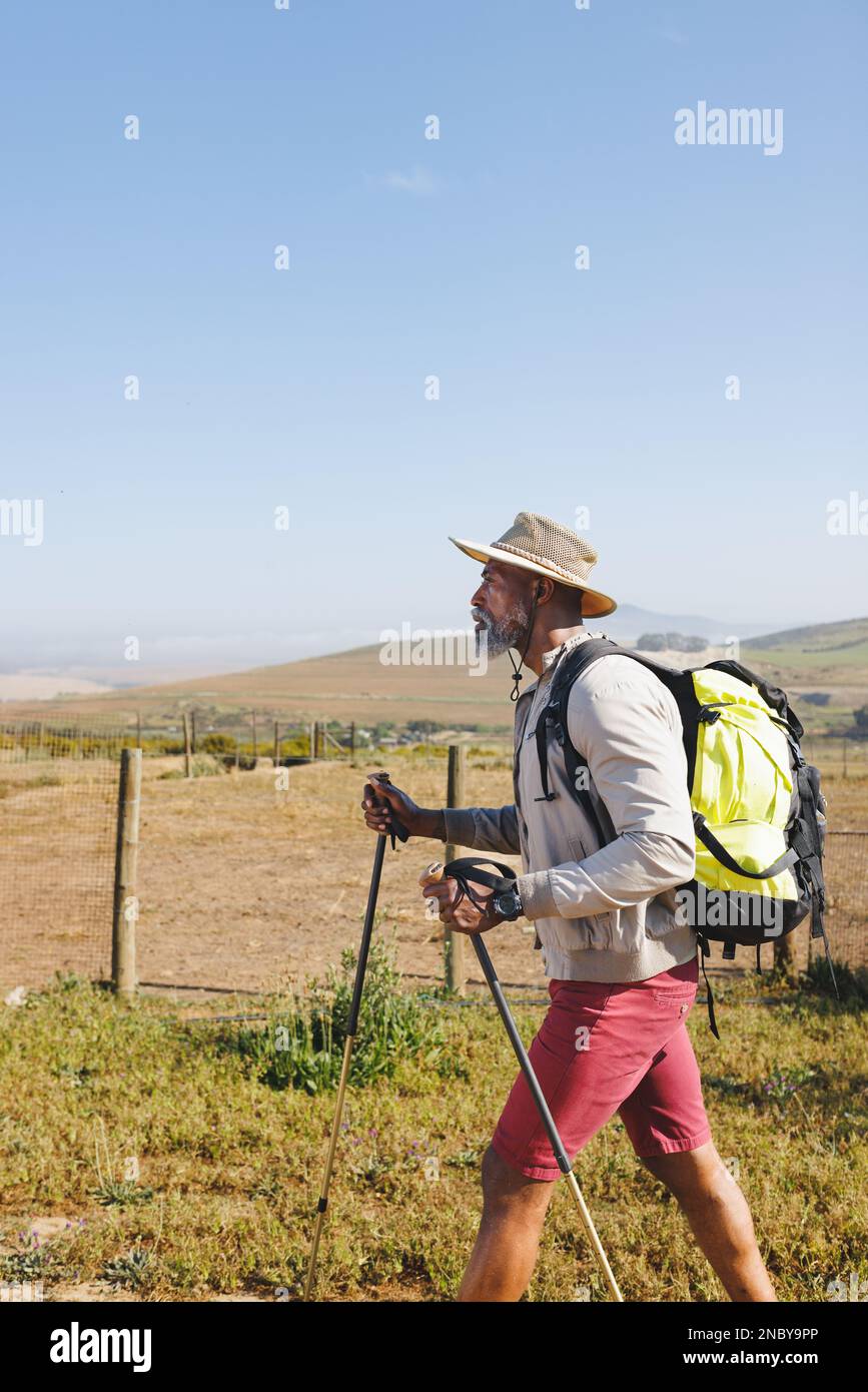 Vue latérale de l'homme senior afro-américain portant un chapeau avec sac à dos et des bâtons de randonnée sur terre. Espace de copie, inchangé, ciel clair, retraite, vacances, s Banque D'Images