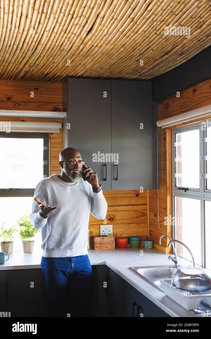 Homme afro-américain de premier plan parlant au-dessus d'un smartphone tout en se tenant près du comptoir de cuisine dans la cabine en rondins. Espace de copie, technologie, chauve, lunettes, unaltere Banque D'Images