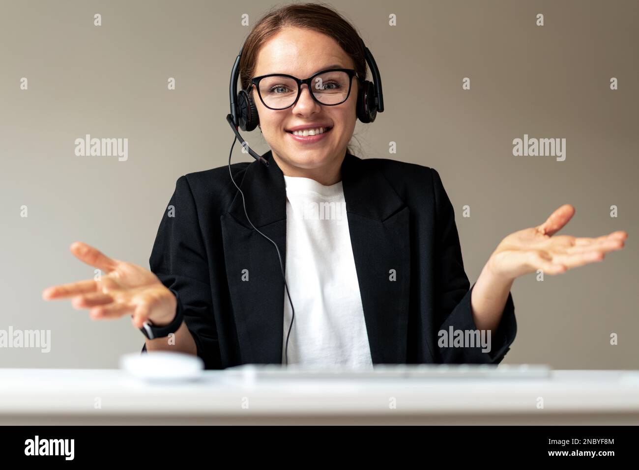 Employée de bureau avec casque et lunettes regardant la webcam sourire et tenant les mains paumes vers le haut. Banque D'Images