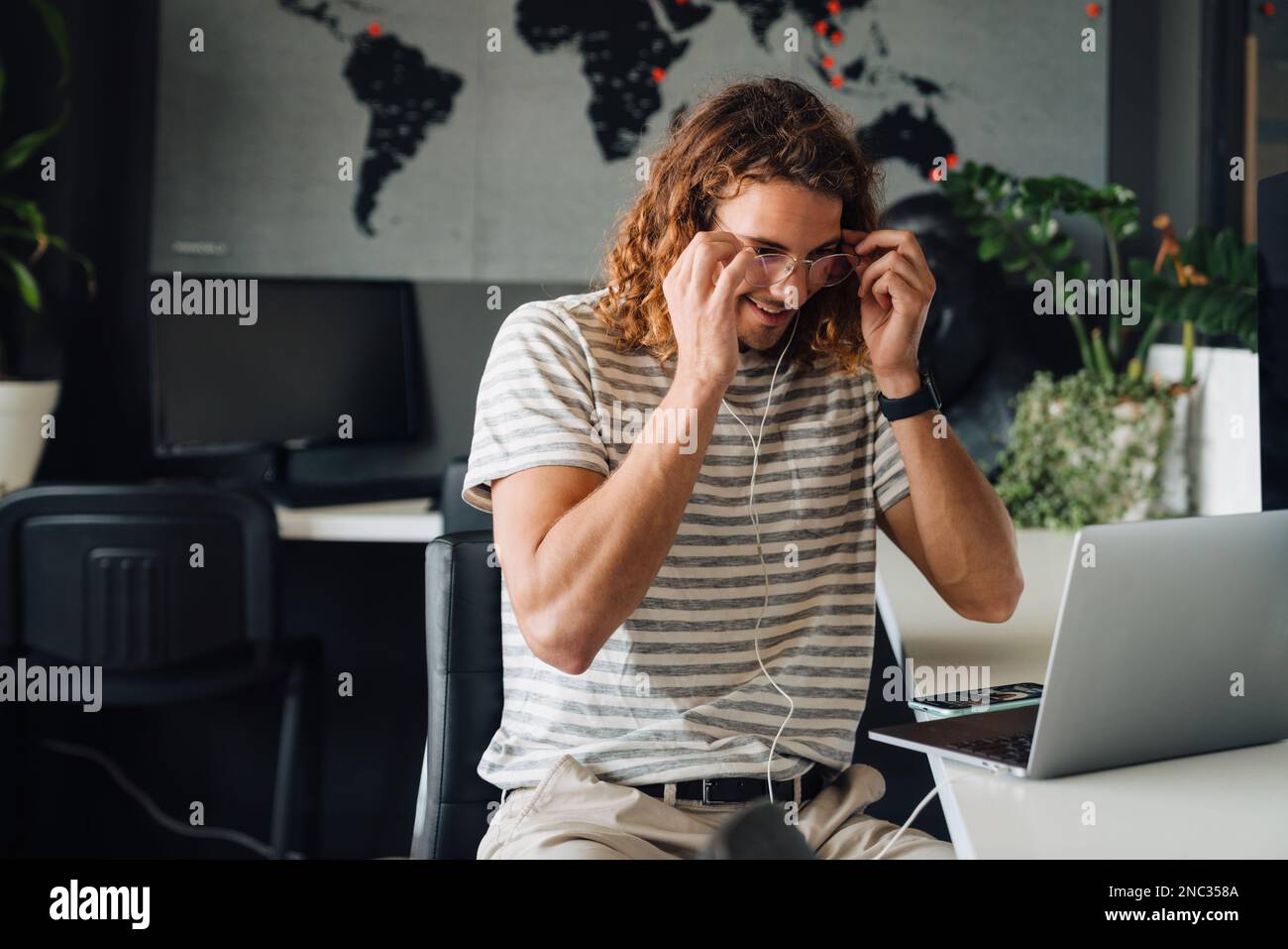Jeune homme d'affaires souriant mettant des lunettes tout en travaillant sur un ordinateur portable assis à une table au bureau Banque D'Images