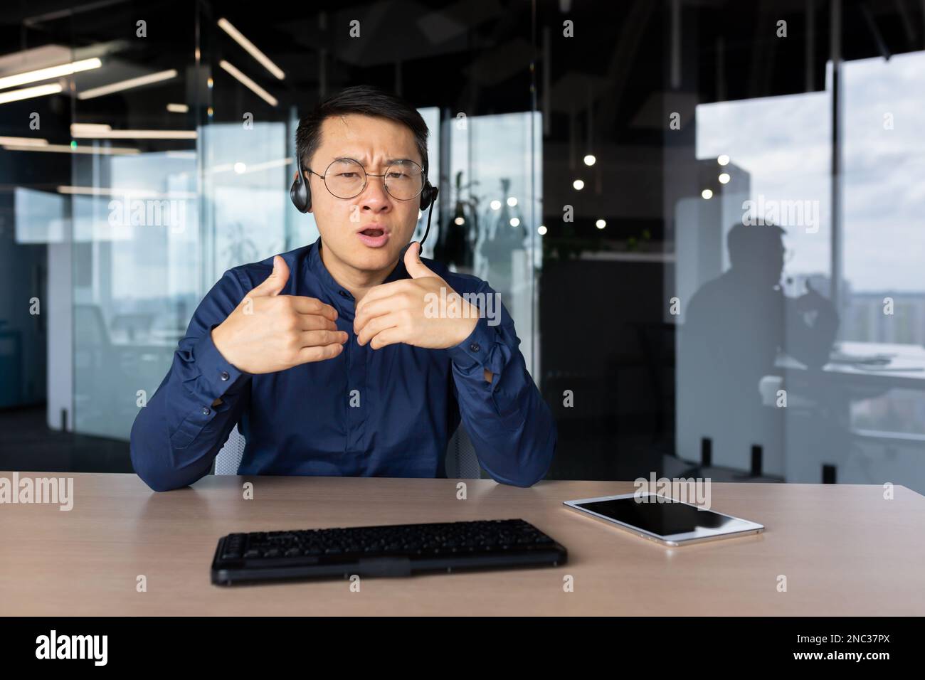 Un jeune homme d'affaires, assistant, consultant est assis au bureau avec enthousiasme et colère lors d'un appel vidéo, au moyen d'un casque. Inquiet et en colère, explique-t-il, en faisant des gestes avec ses mains. Banque D'Images