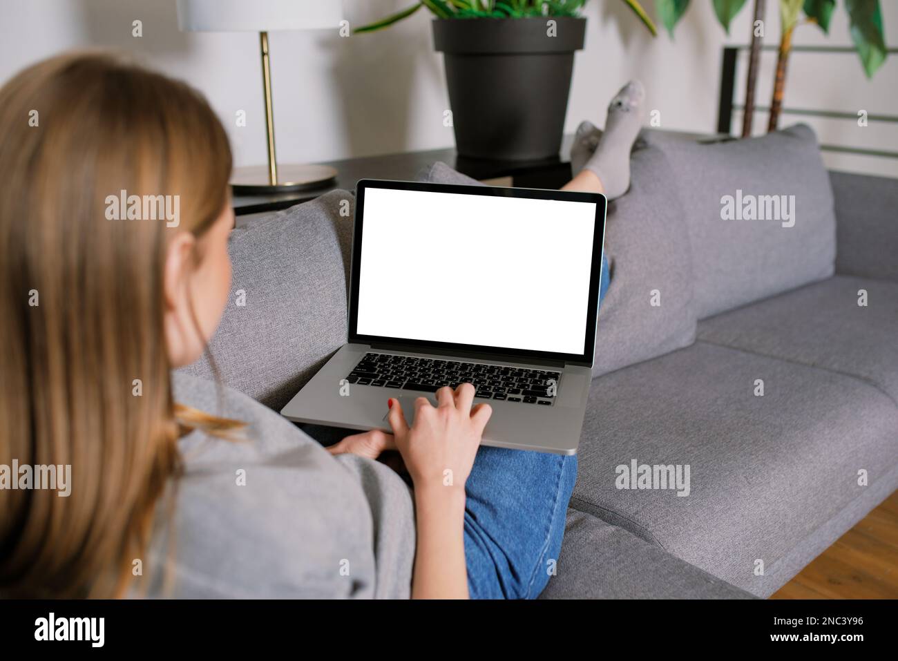Jeune femme blonde décontractée méconnaissable dans un salon moderne, couchée sur un canapé avec des jambes croisées, travaillant avec écran vierge ordinateur portable maquette Banque D'Images