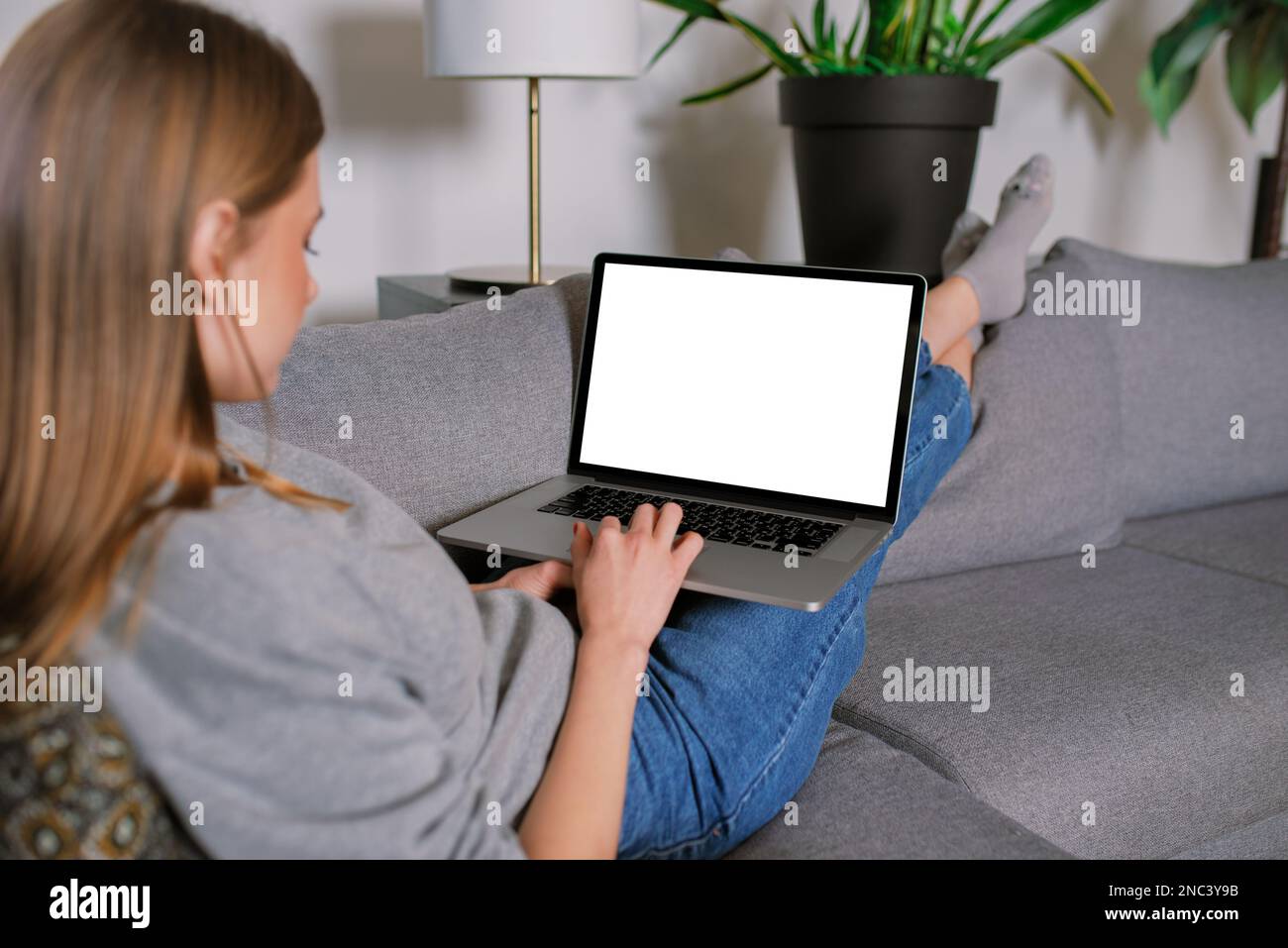 Jeune femme blonde décontractée méconnaissable dans un salon moderne, couchée sur un canapé avec des jambes croisées, travaillant avec écran vierge ordinateur portable maquette Banque D'Images