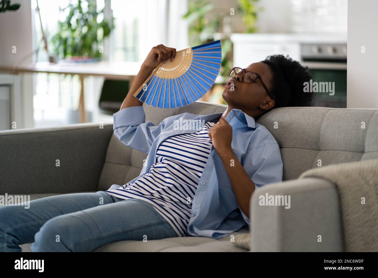 Une femme afro-américaine fatiguée et épuisée s'assoit sur un canapé en utilisant des fans de papier souffrant de stuffess Banque D'Images