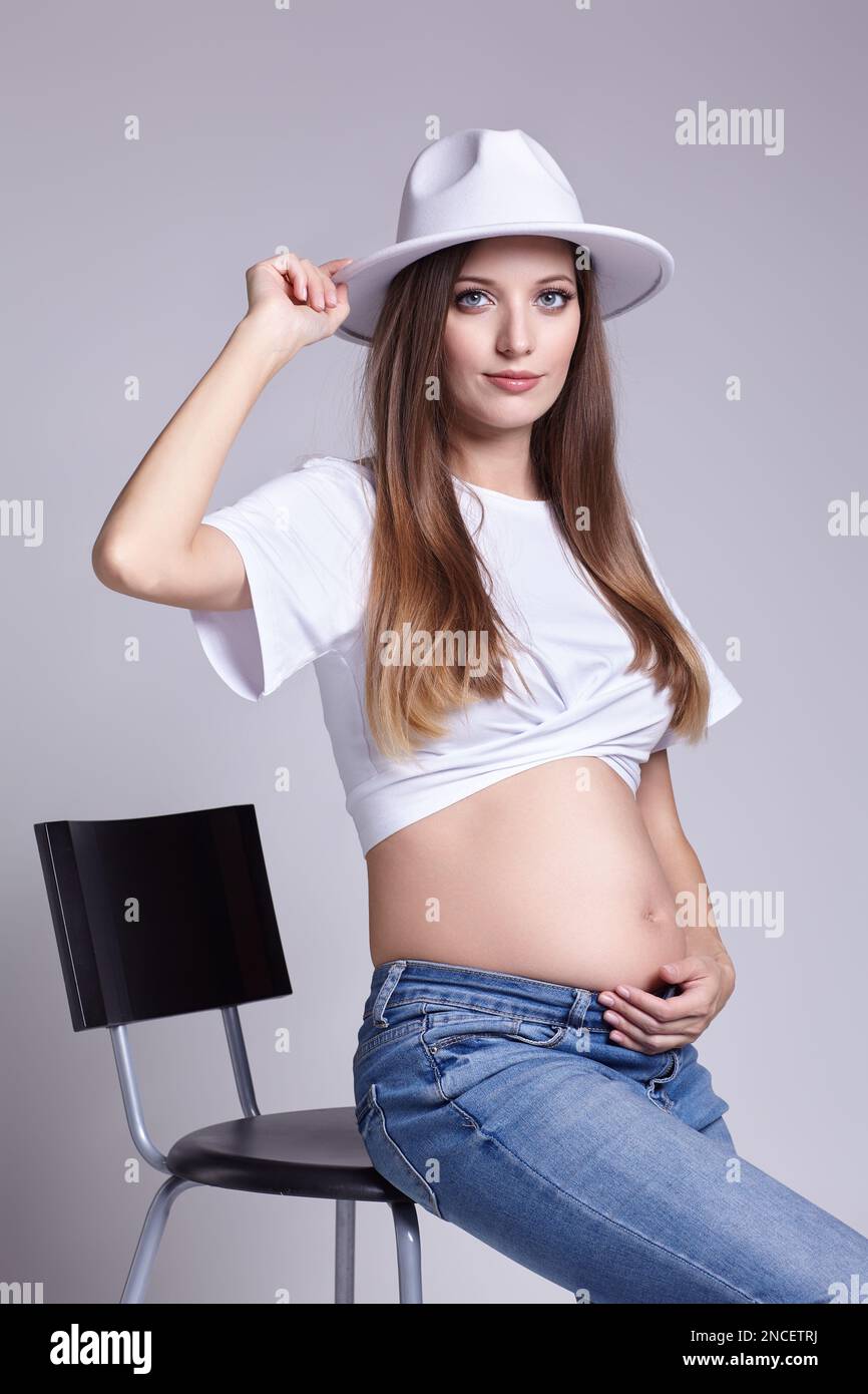 Jeune femme enceinte en t-shirt blanc, et un Jean s'assoit sur un tabouret  de bar. Femelle avec le ventre exposé. 5th mois de grossesse Photo Stock -  Alamy