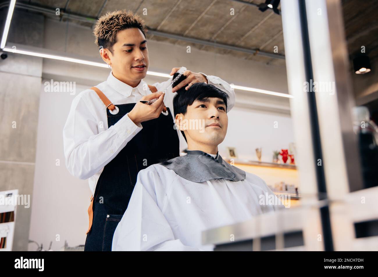 Joyeux coiffeur de jeune asiatique souriant portant un tablier dans moderne salon tenant la brosse et les ciseaux de couper les cheveux de client Banque D'Images