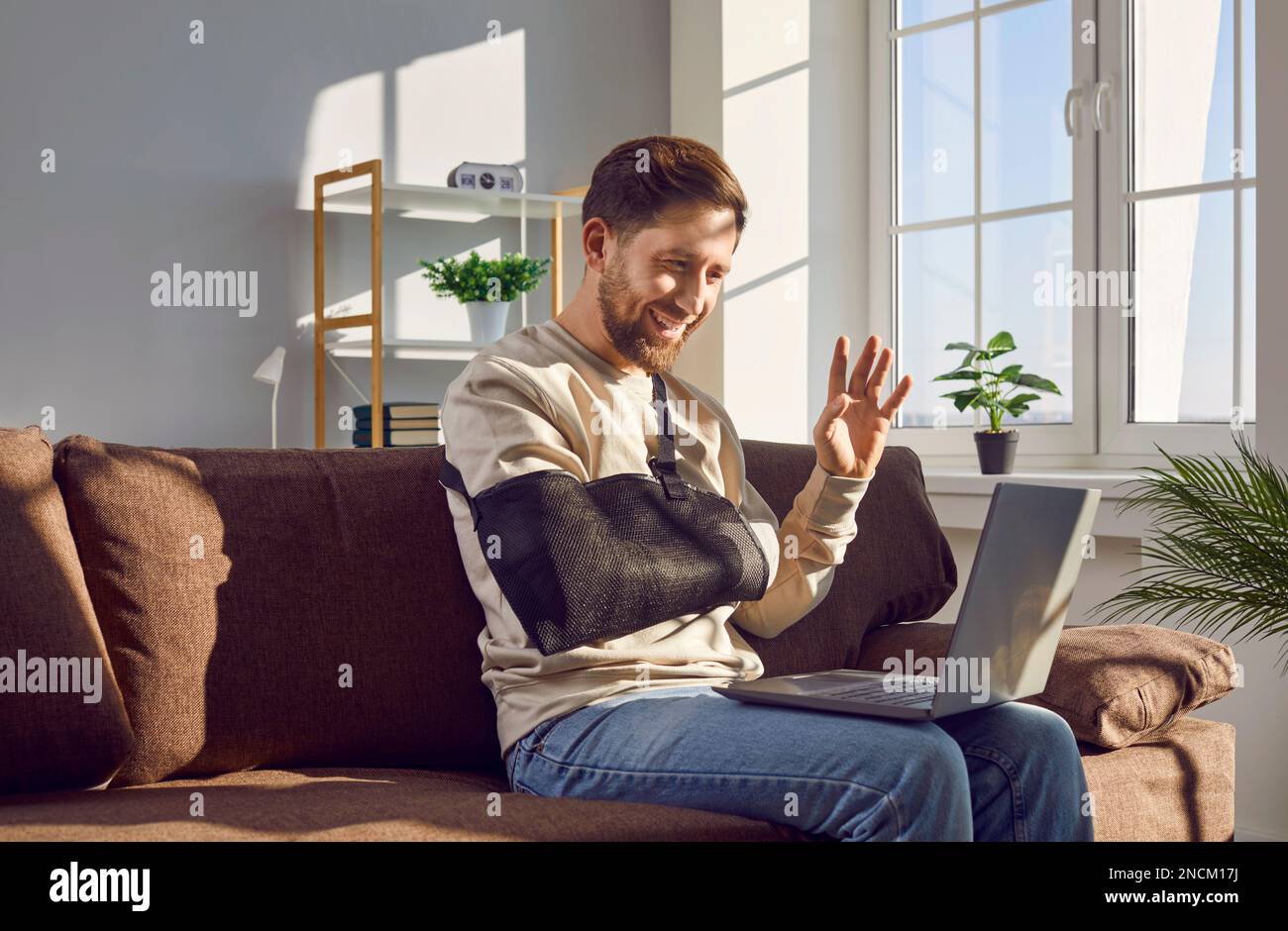 Un jeune homme souriant avec un bras cassé qui effectue un appel vidéo à l'aide d'un ordinateur portable Banque D'Images