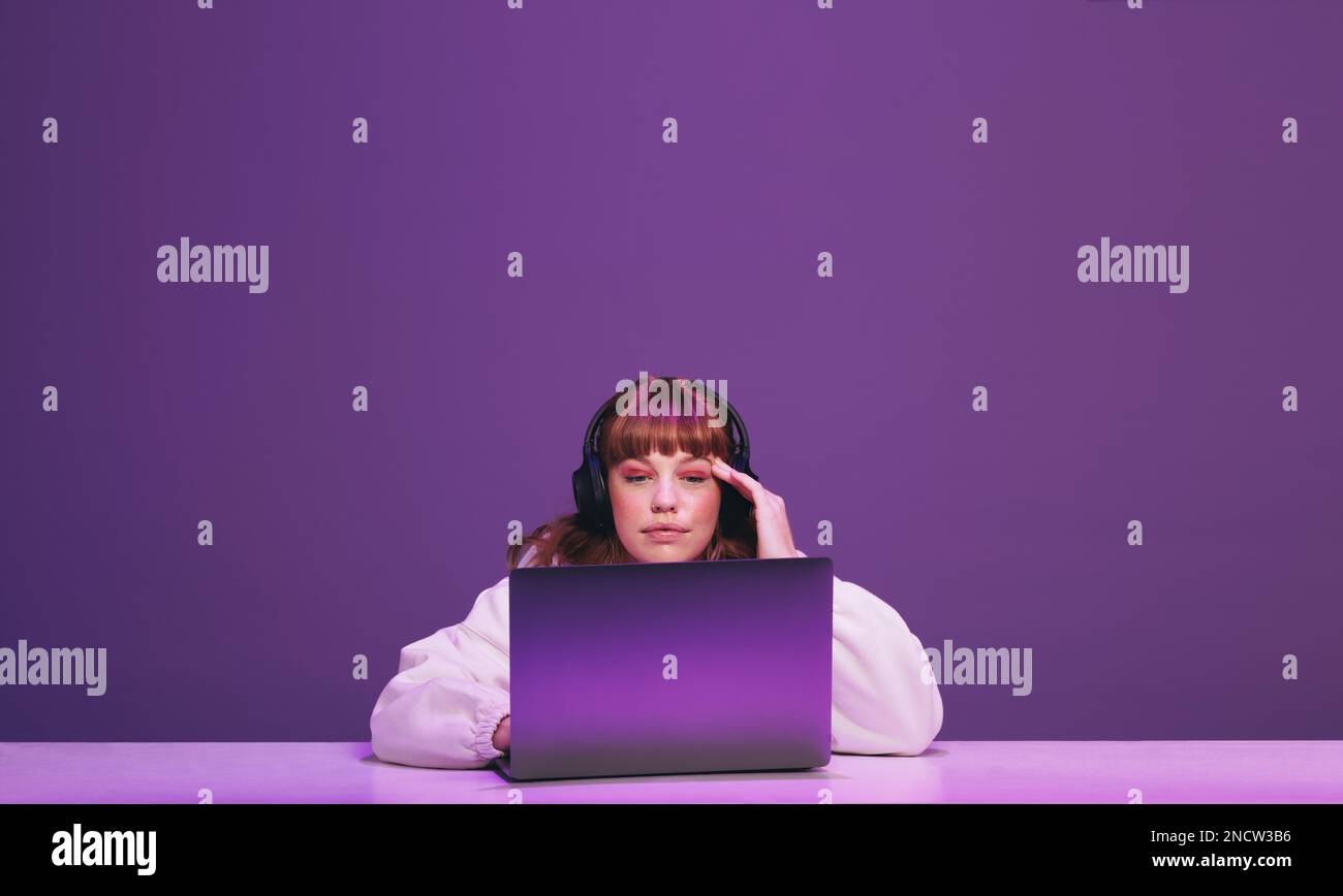 Femme aux cheveux de gingembre en pensant tout en jouant à un jeu vidéo en ligne sur un ordinateur portable. Jeune joueur utilisant un service de streaming interactif dans un studio. Banque D'Images