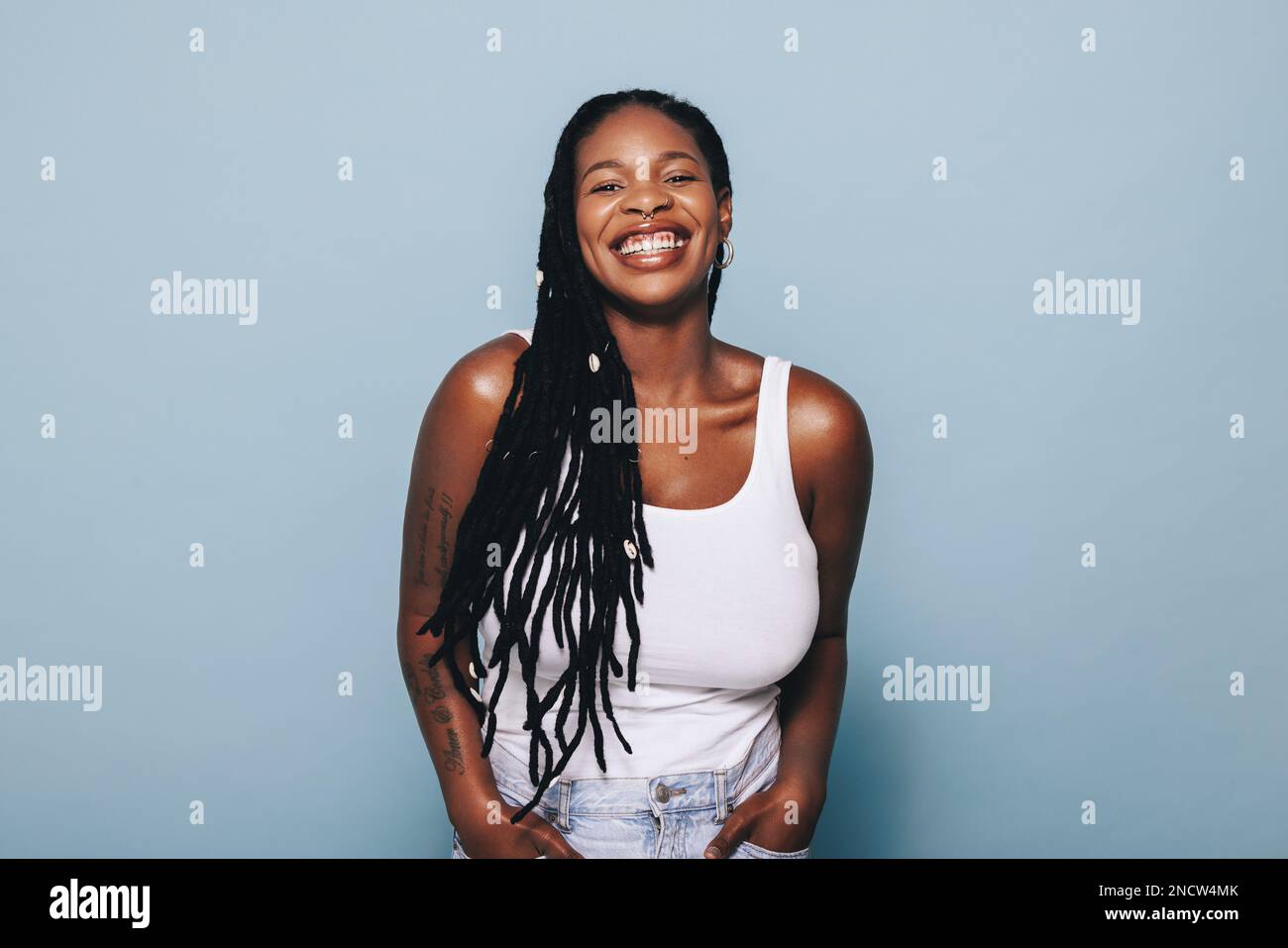 Une jeune femme heureuse souriant à l'appareil photo tout en se tenant sur un fond bleu dans des vêtements décontractés. Jeune femme noire élégante portant des tatouages de bras et Banque D'Images
