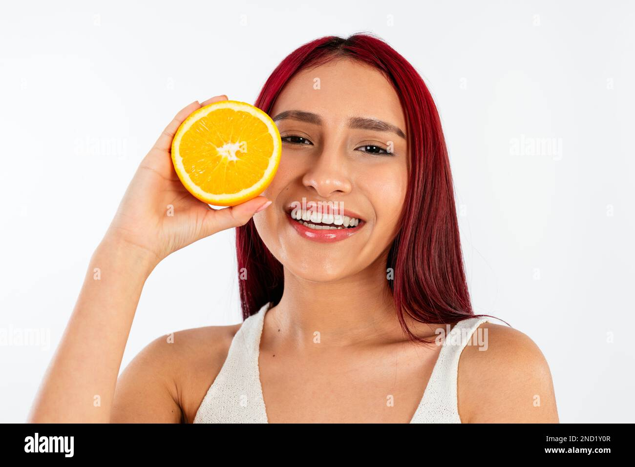 Jeune femme heureuse avec des cheveux rouge vif souriant tout en tenant des moitiés d'oranges contre fond blanc regardant l'appareil photo Banque D'Images