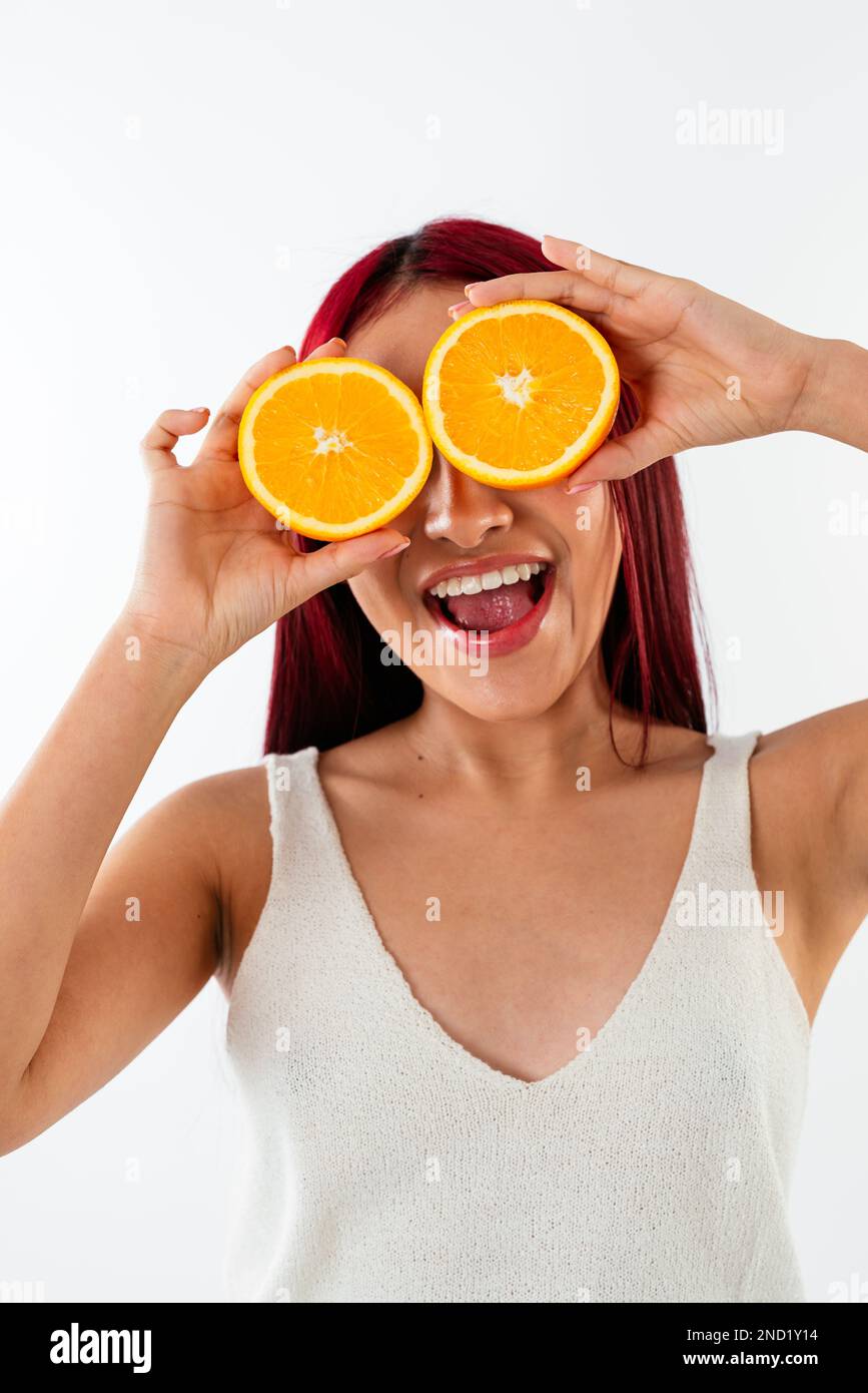 Jeune femme heureuse avec des cheveux rouge vif souriant et cachant les yeux derrière des moitiés d'oranges sur fond blanc Banque D'Images