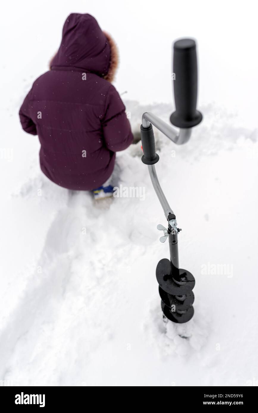 Exercice de glace pour la pêche d'hiver avec des vis se tient devant la fille assise et des trous de pêche sur la glace de la rivière dans la neige blanche. Banque D'Images