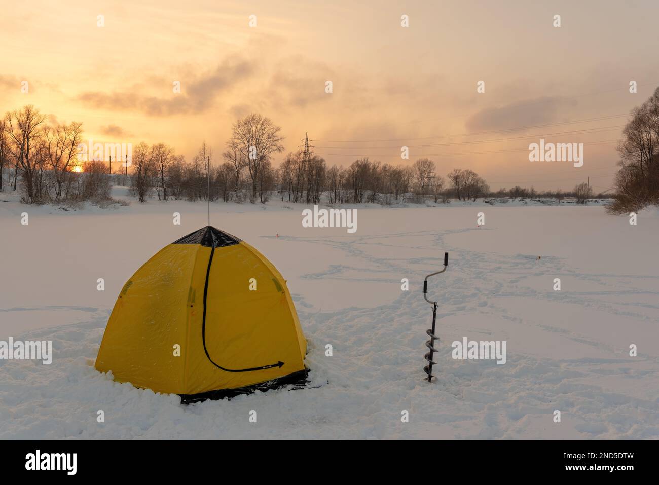 Tente jaune d'hiver avec vis à glace près d'un pêcheur se tenant sur la glace d'une rivière sibérienne sur fond de coucher de soleil derrière la forêt. Banque D'Images