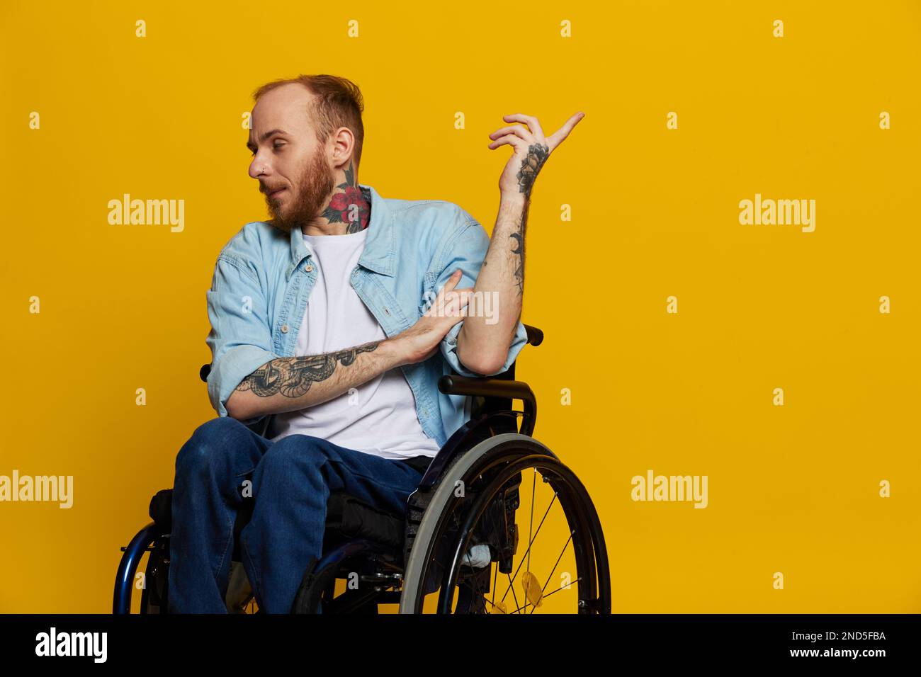 Un homme en fauteuil roulant a des problèmes avec le système musculo-squelettique regarde la caméra montre un doigt dessus, avec des tatouages sur ses mains assis sur un jaune Banque D'Images