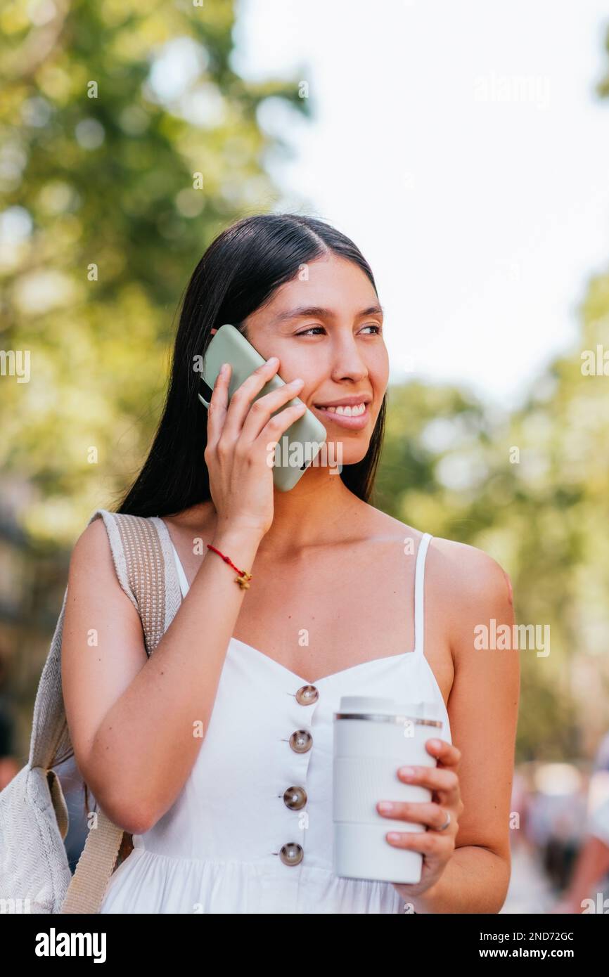 Bonne femme latino-américaine avec une tasse de café à emporter sans gaspillage souriant et regardant loin tout en parlant sur téléphone portable sur un fond flou de parc en été à Barcelone, Espagne Banque D'Images