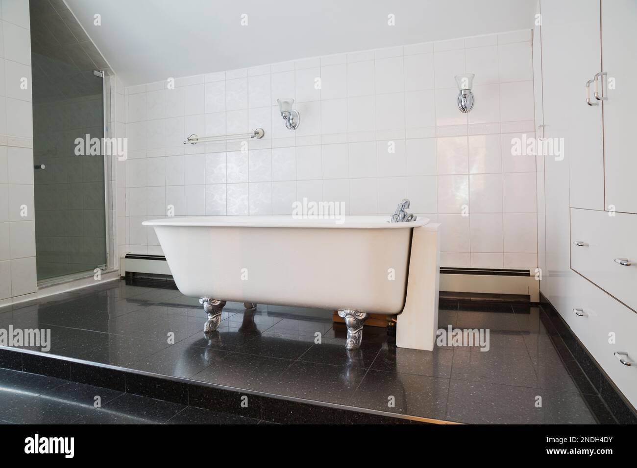 Baignoire à griffes en fonte antique blanche sur plate-forme surélevée en granit noir dans la salle de bains principale avec armoires et tiroirs blancs dans la vieille maison de 1900. Banque D'Images