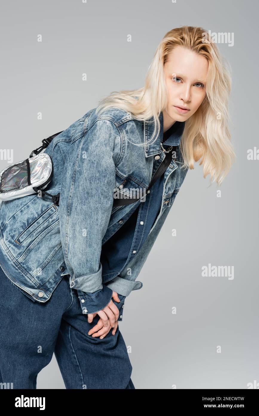 jeune blond modèle dans une élégante tenue en denim et sac de ceinture tendance posant isolé sur gris, image de stock Banque D'Images