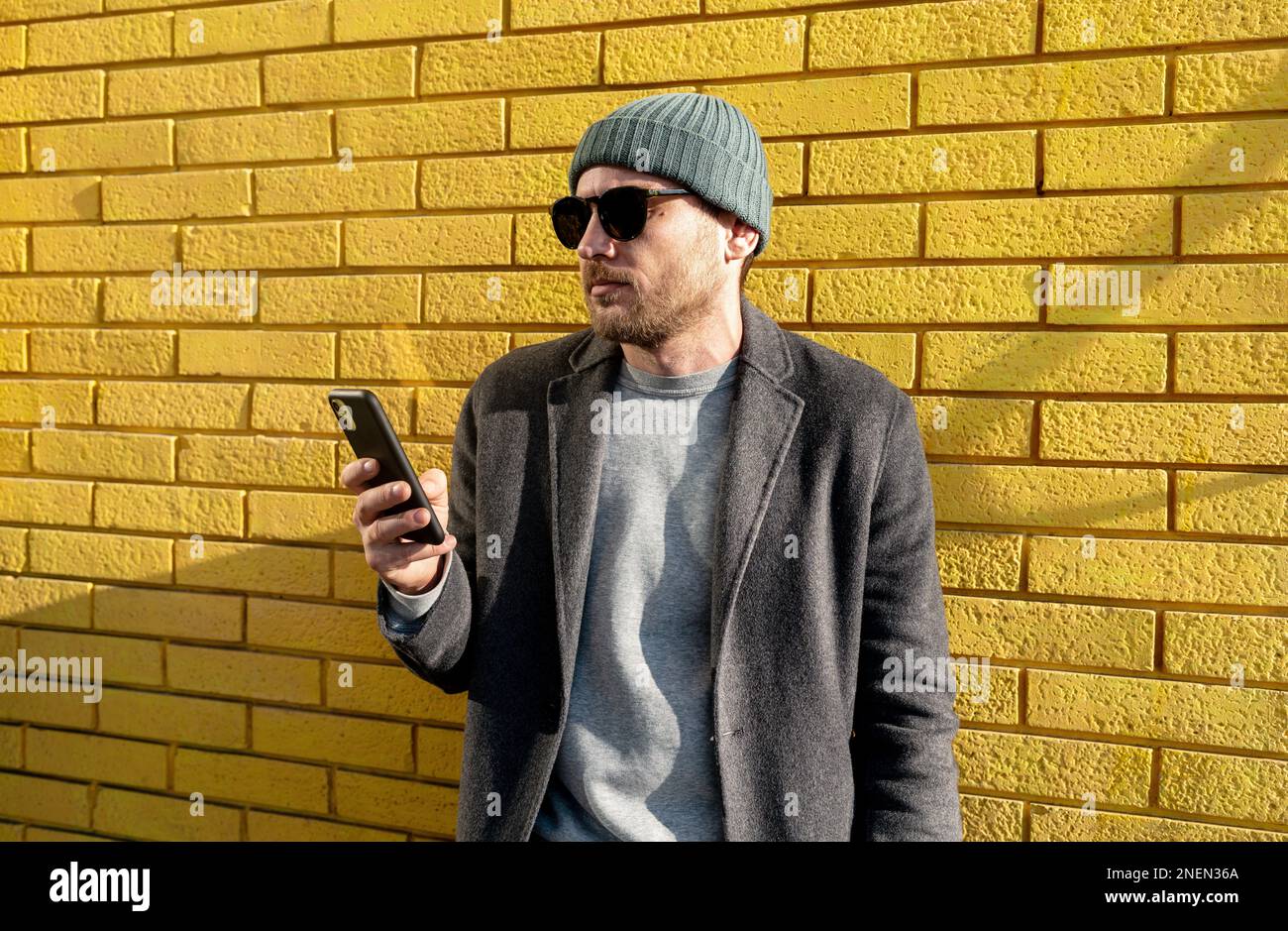 Homme urbain élégant dans un chapeau avec des lunettes de soleil se tenant contre le mur de brique jaune et utilisant son téléphone cellulaire. Banque D'Images