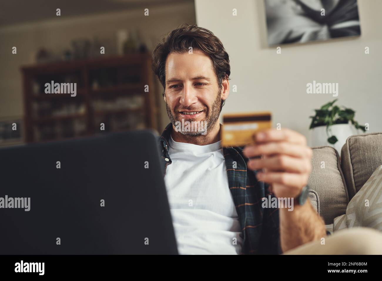 À la recherche de promotions en ligne. un beau jeune homme utilisant son ordinateur portable et sa carte de crédit tout en se relaxant sur un canapé à la maison. Banque D'Images