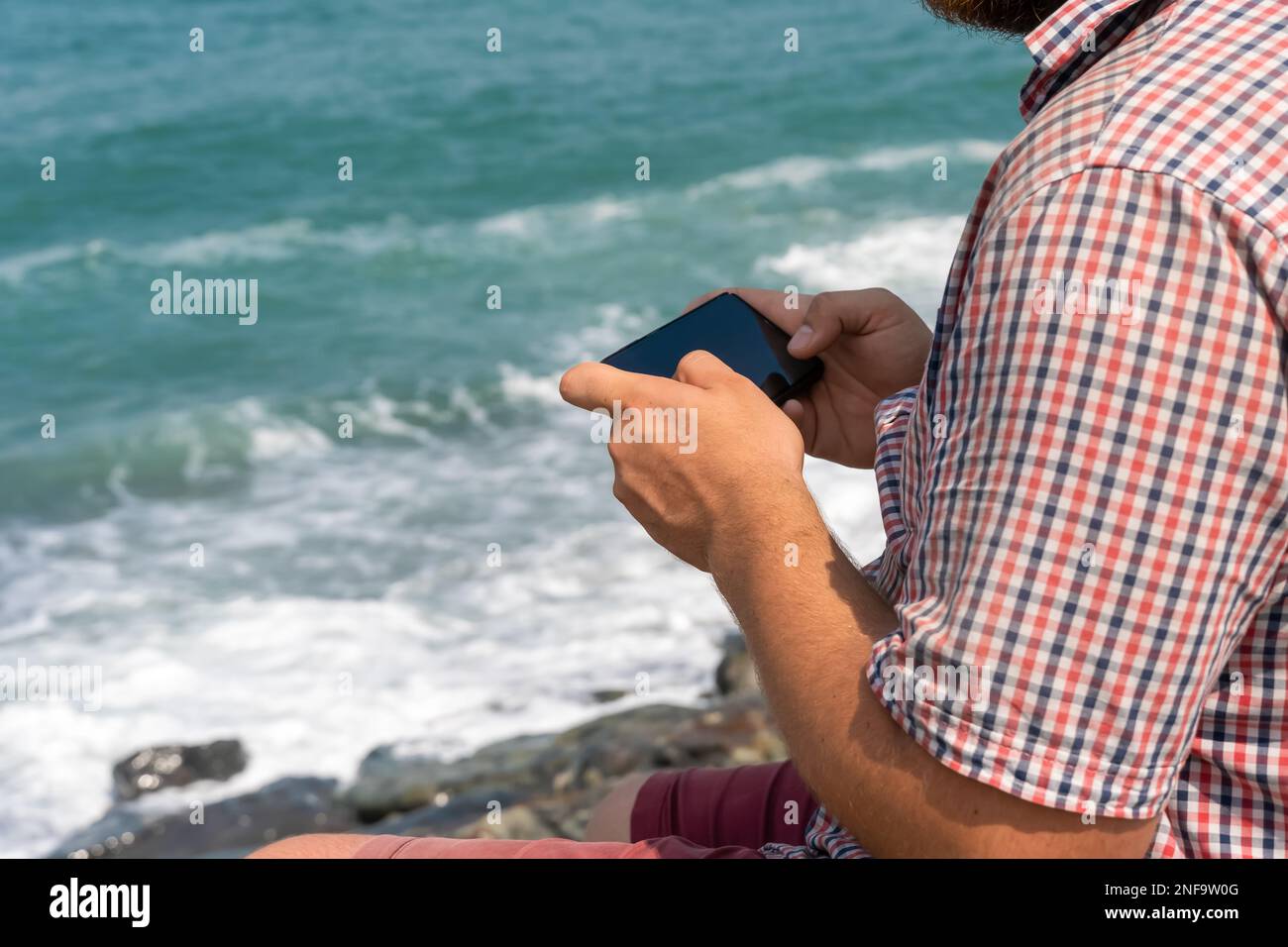 Homme dans une chemise à carreaux assis sur le bord de mer par temps ensoleillé et tenant un téléphone portable dans ses mains Banque D'Images