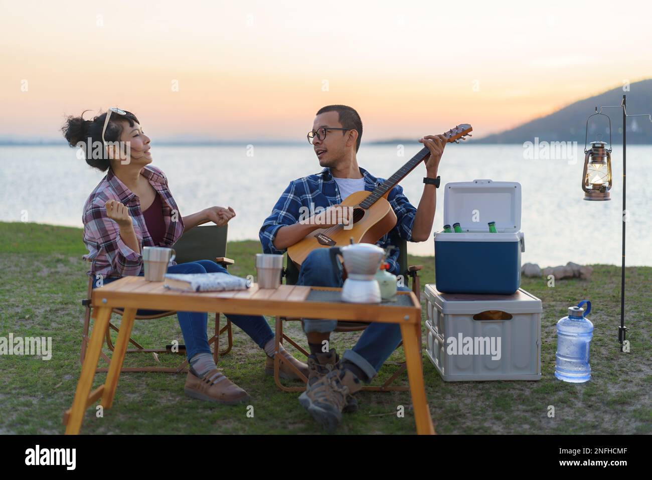 Couple asiatique chantant et jouant de la guitare dans leur zone de camping avec lac en arrière-plan pendant le coucher du soleil. Banque D'Images